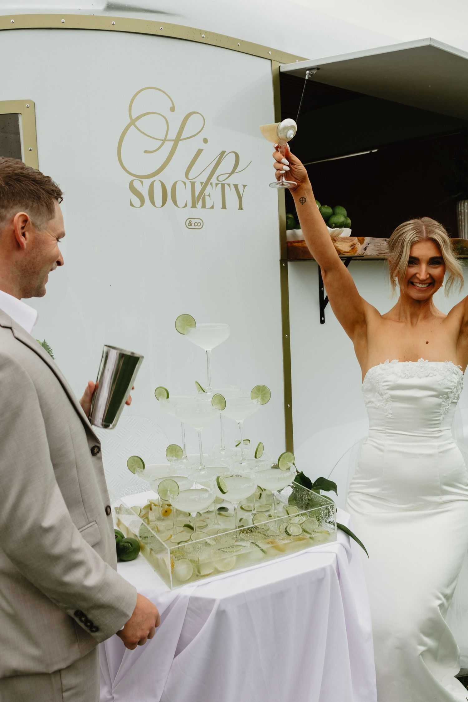 A bride raises a champagne glass at a Sip Society bar beside her groom at a wedding reception.