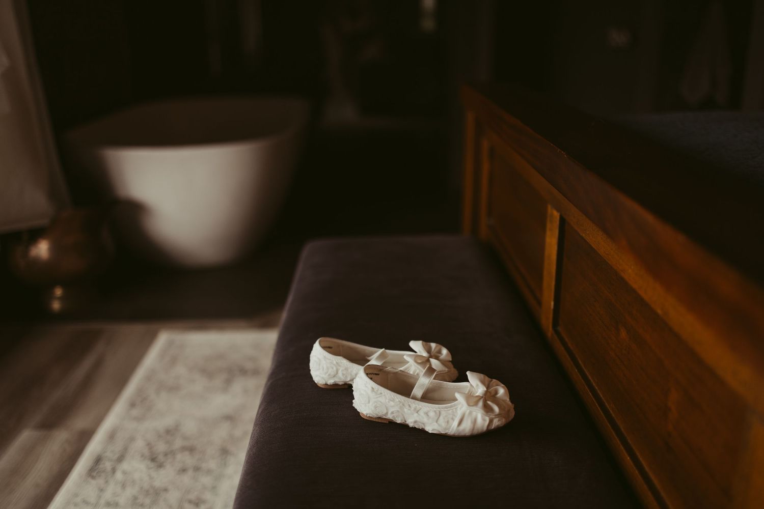 White bridal ballet flats with bow detail resting on a dark upholstered bench in dim room.