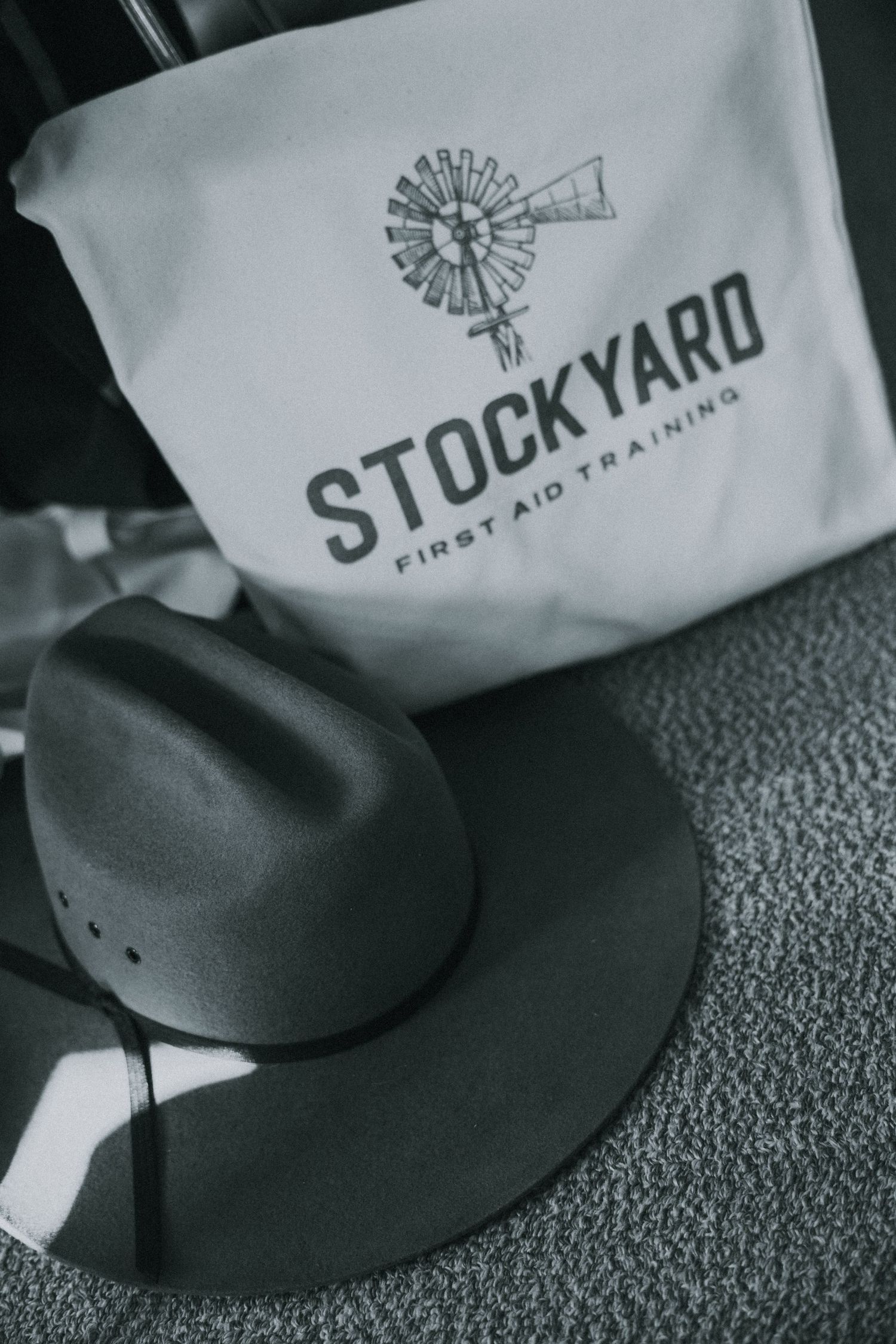 Black and white photo of a Stockyard First Aid Training bag beside a wide-brim hat on a textured surface.