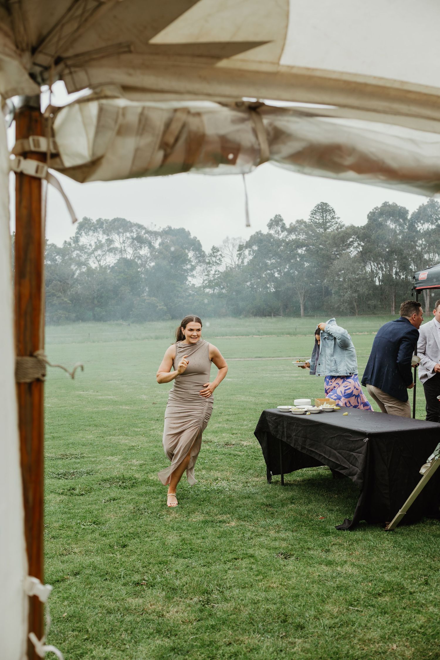 Guest in beige dress walking barefoot across green grass toward a rustic wedding marquee.