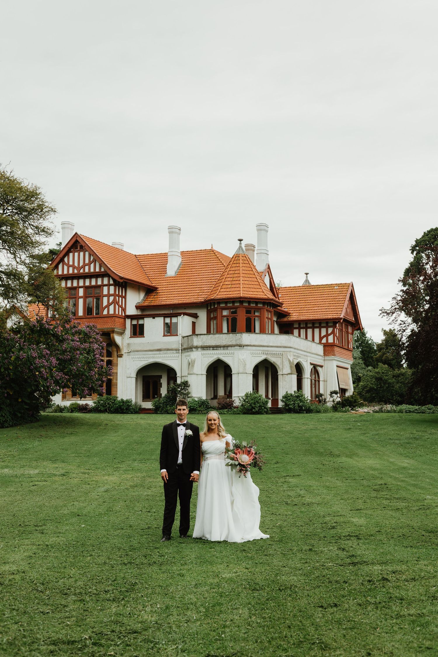 Bride and groom pose before grand Tudor-style mansion with orange roof on green lawn.