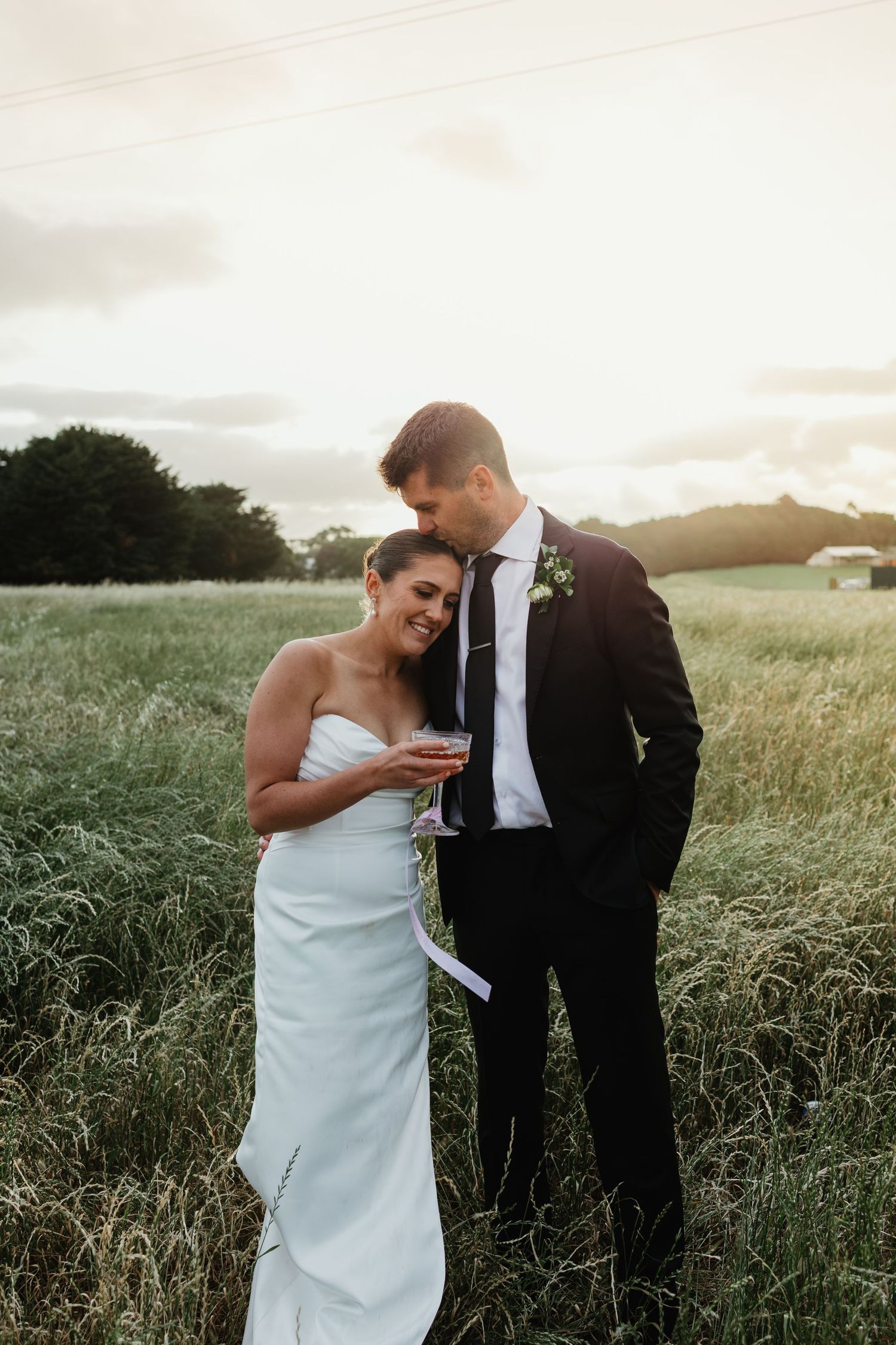 Bride and groom share a romantic moment in a golden wheat field at sunset, bride holding a glass of wine.