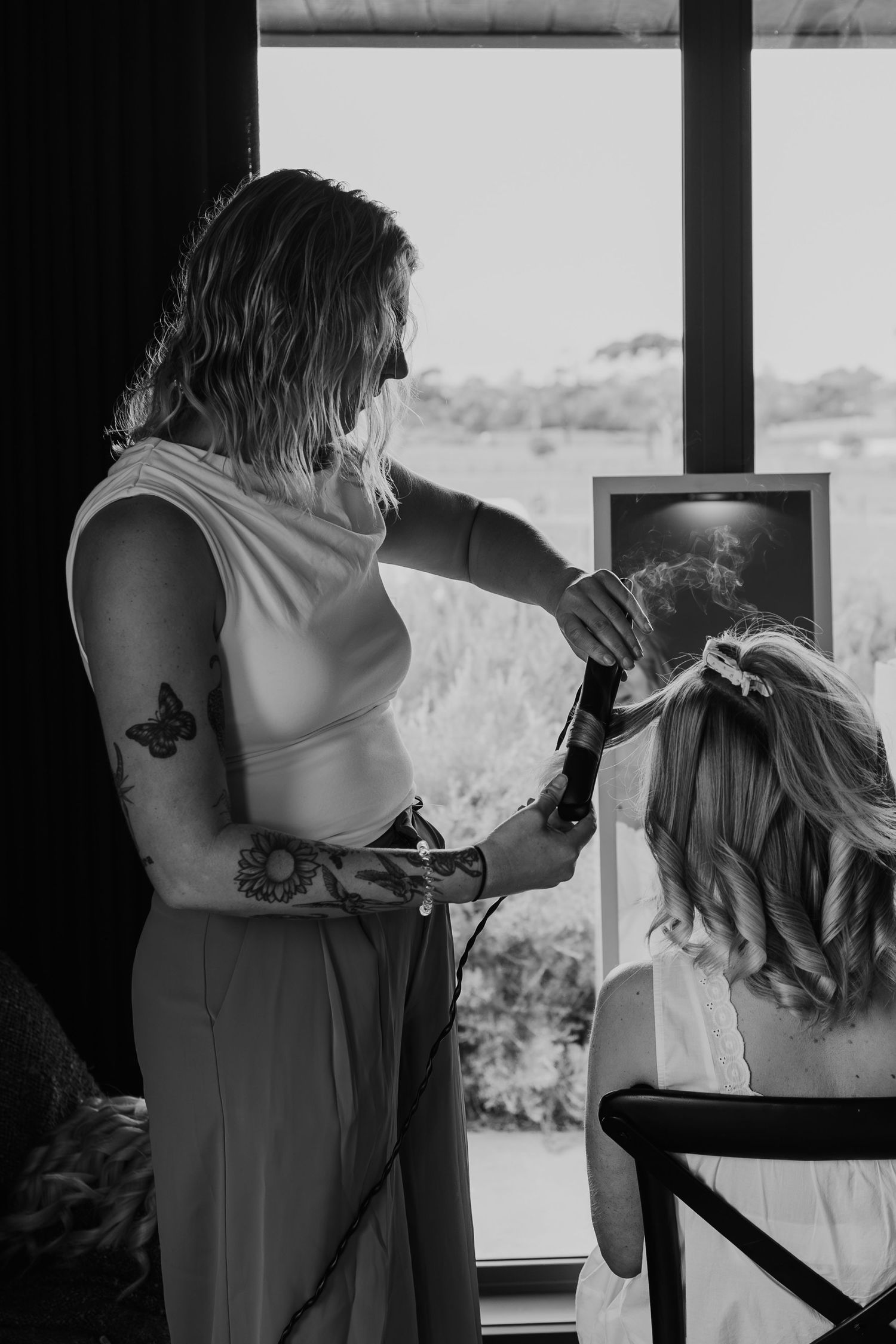 Hair stylist using curling iron on blonde bride's hair in black and white wedding preparation photo.