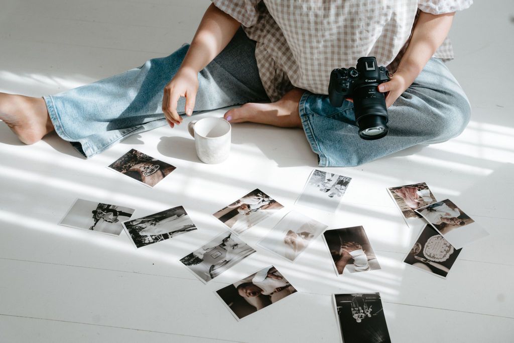 Photographer sitting cross-legged on white floor reviewing printed photos with a DSLR camera in hand.