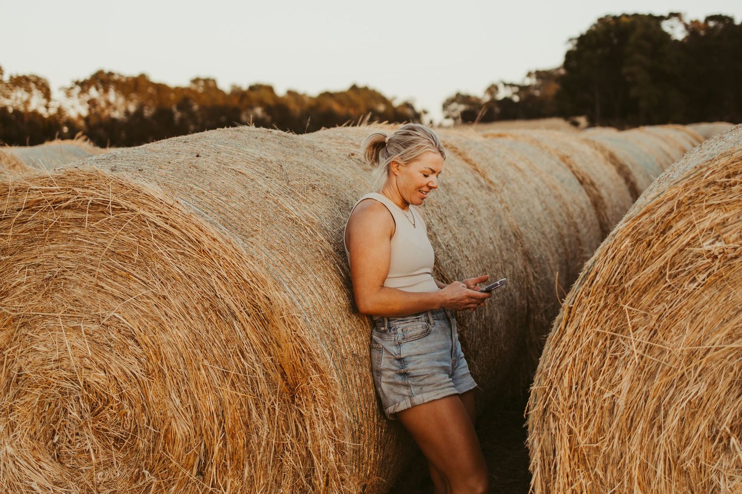 Young woman smiling at her phone while leaning against large hay bales in a golden farm field at sunset.