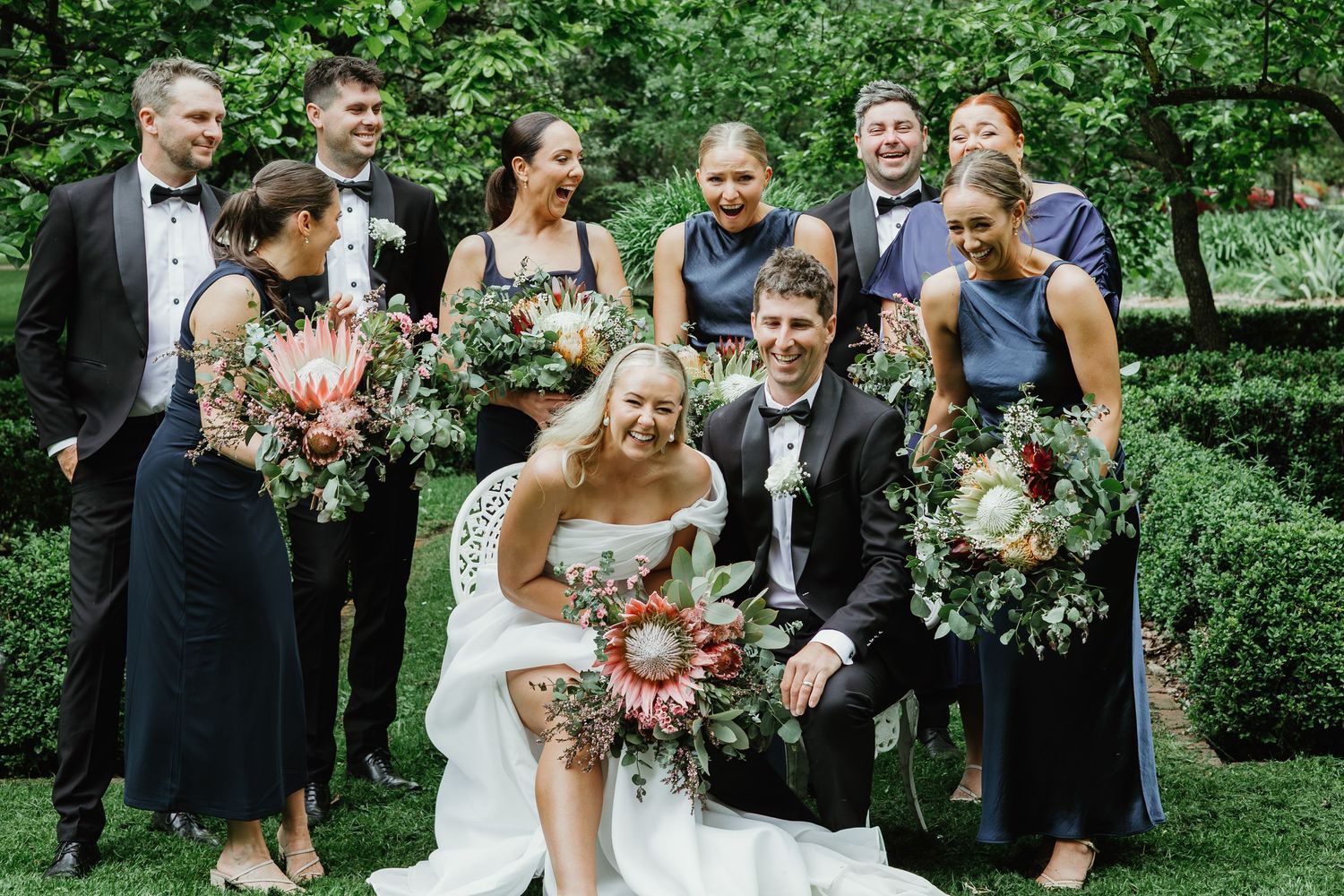 Joyful wedding party with protea bouquets gathered around bride and groom in a lush green garden setting.