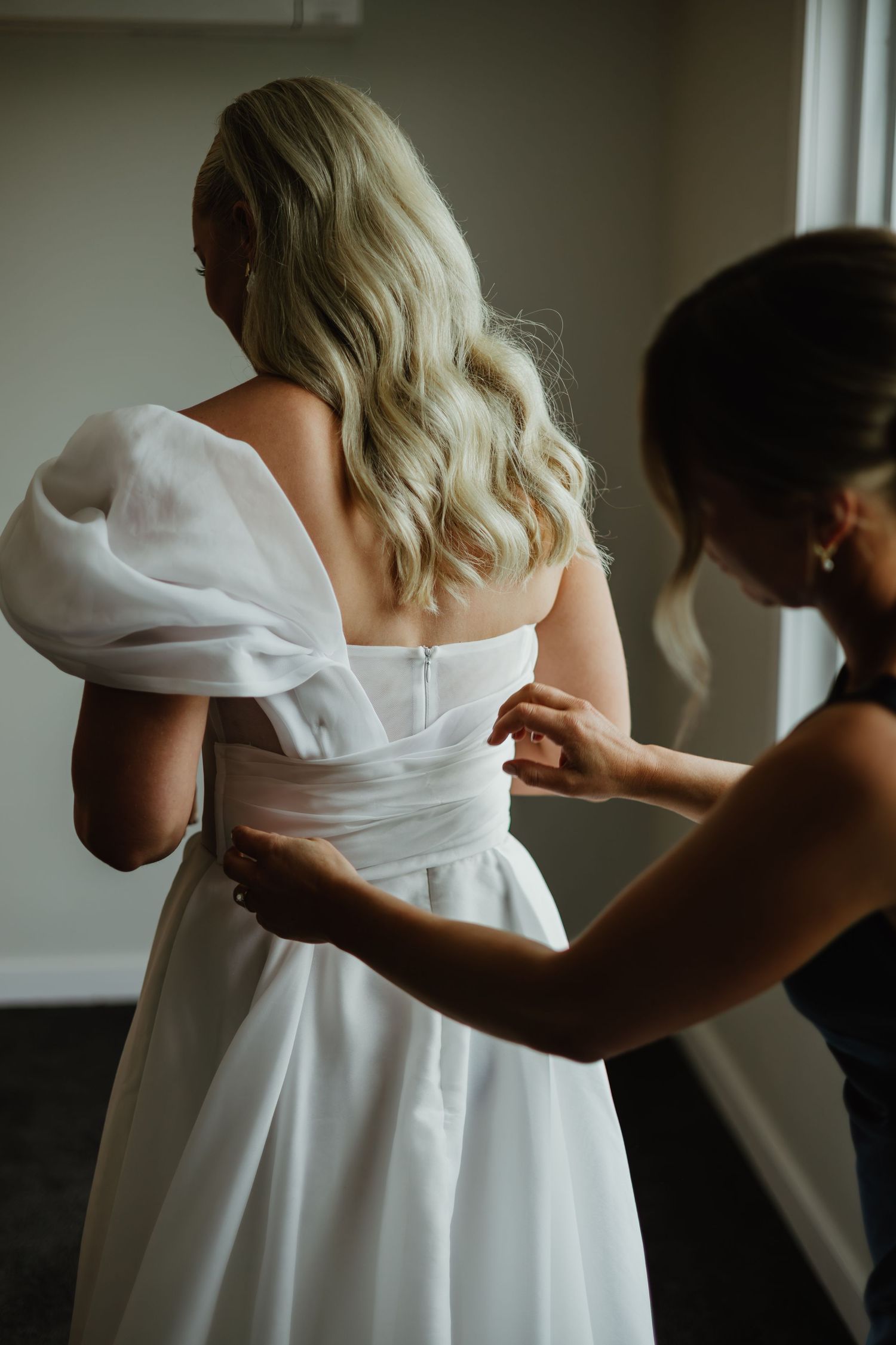 Bridesmaid fastening buttons on back of brides white off-shoulder ballgown wedding dress.