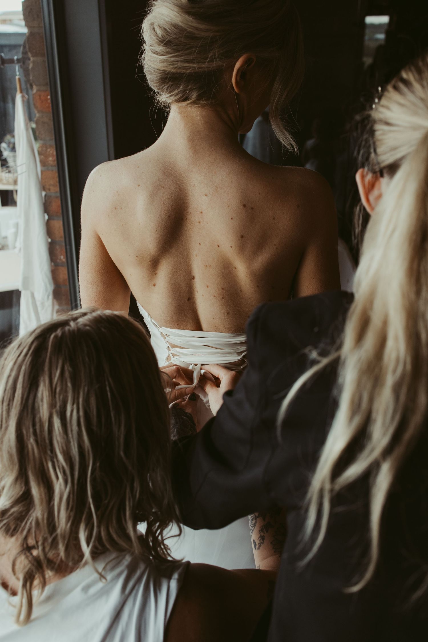 Bridesmaids helping bride fasten her white wedding dress from behind near a window.