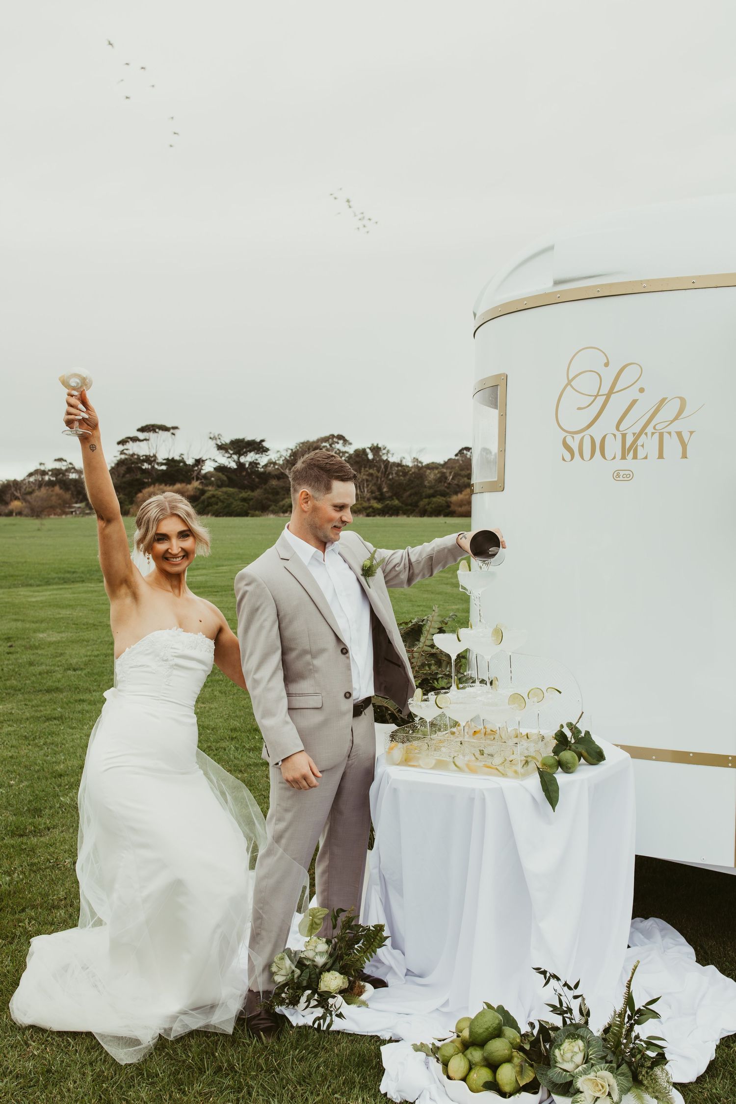 Bride and groom celebrating at a Sip Society champagne tower on a green outdoor wedding venue.
