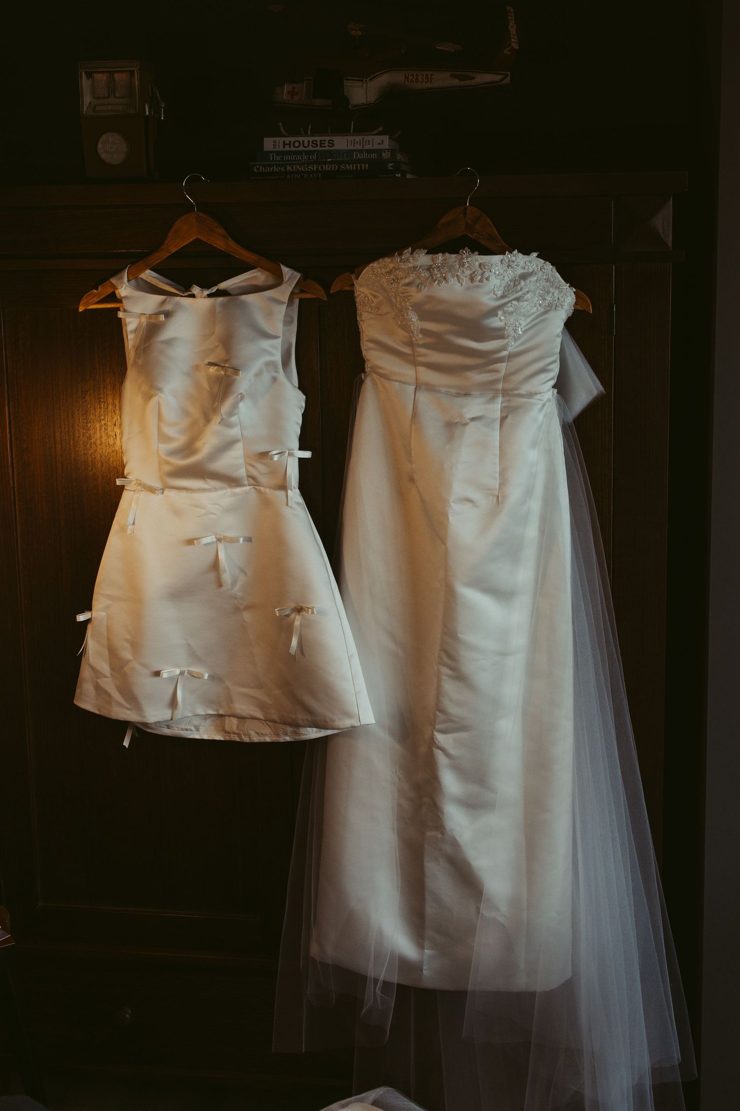 Two white wedding dresses hanging on wooden hangers, including a flower girl dress with bows and a strapless bridal gown with veil.