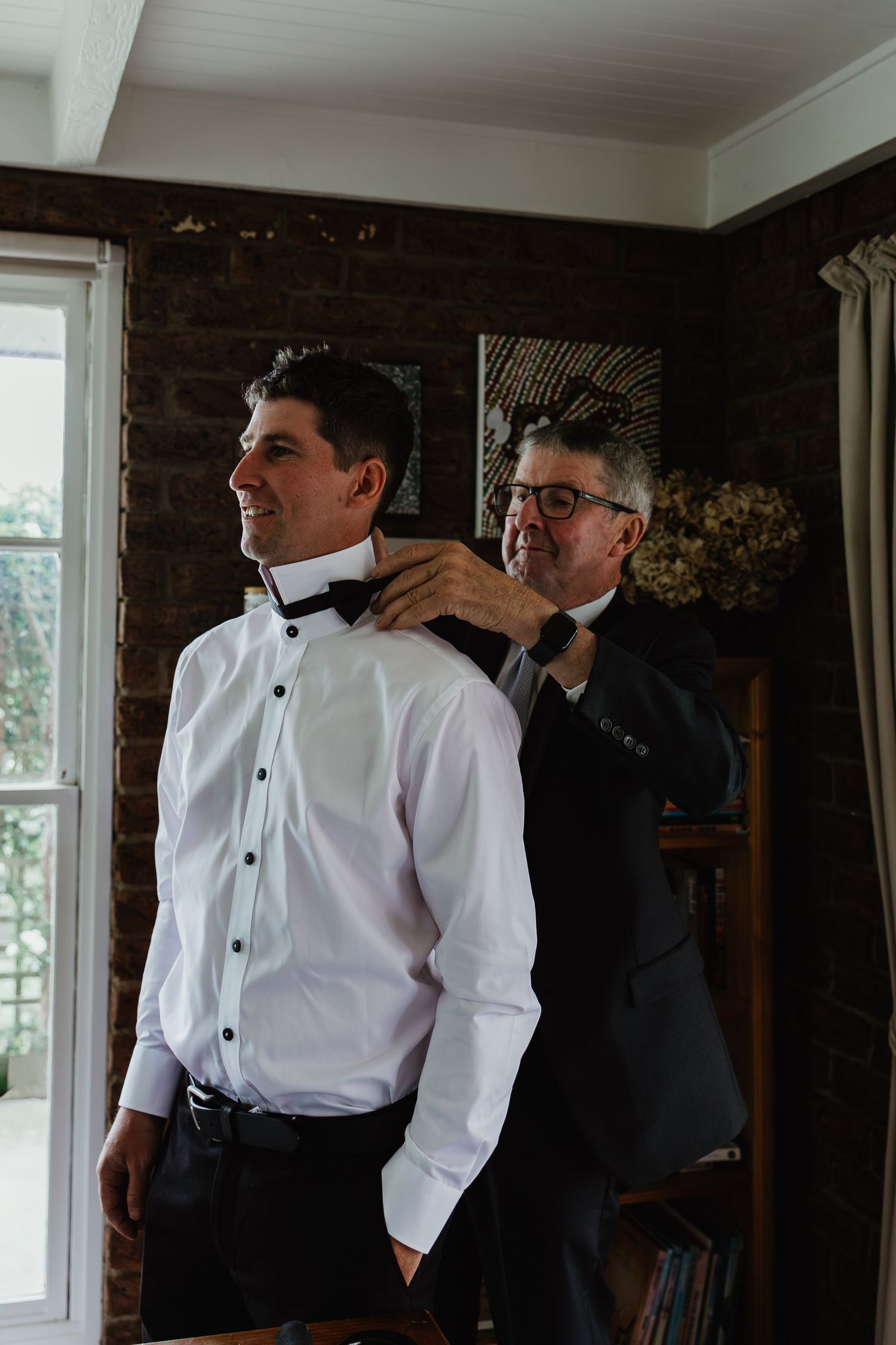 Older man helps groom adjust bow tie in warmly lit room before wedding ceremony.