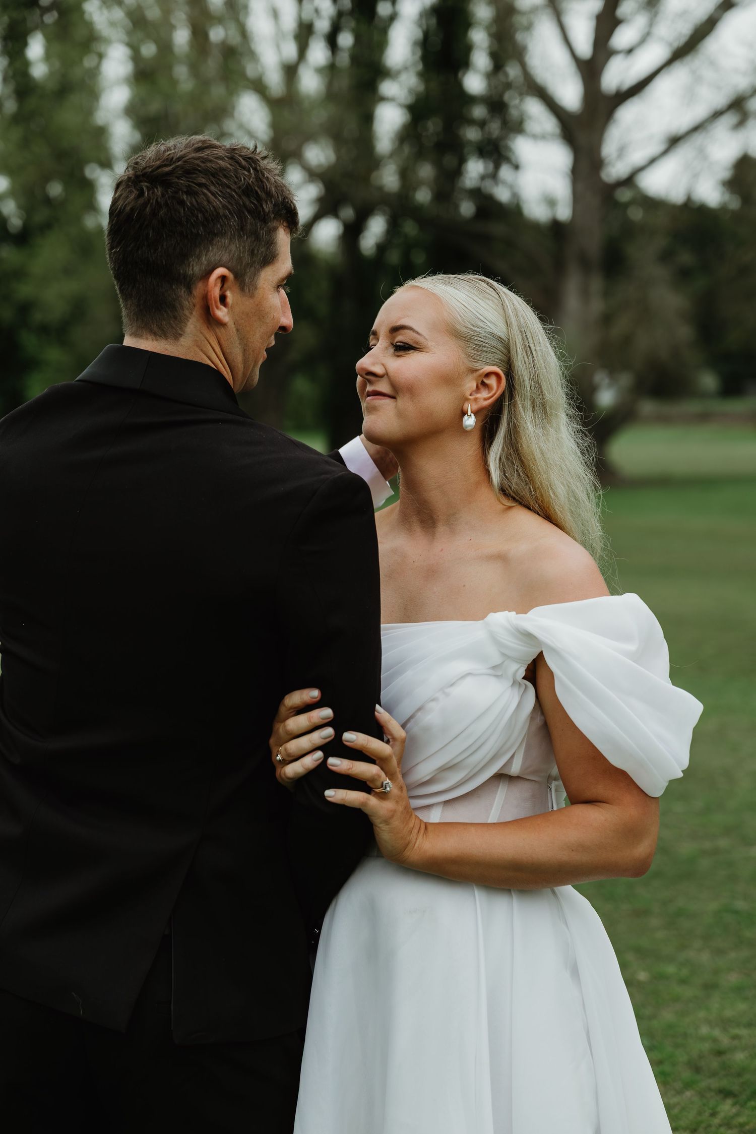 Bride in off-shoulder white gown smiling at groom in black suit during outdoor wedding portraits.