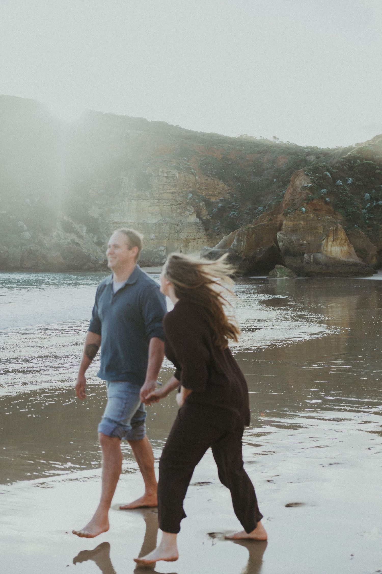A couple walks hand in hand along a misty shoreline with dramatic rocky cliffs behind them.
