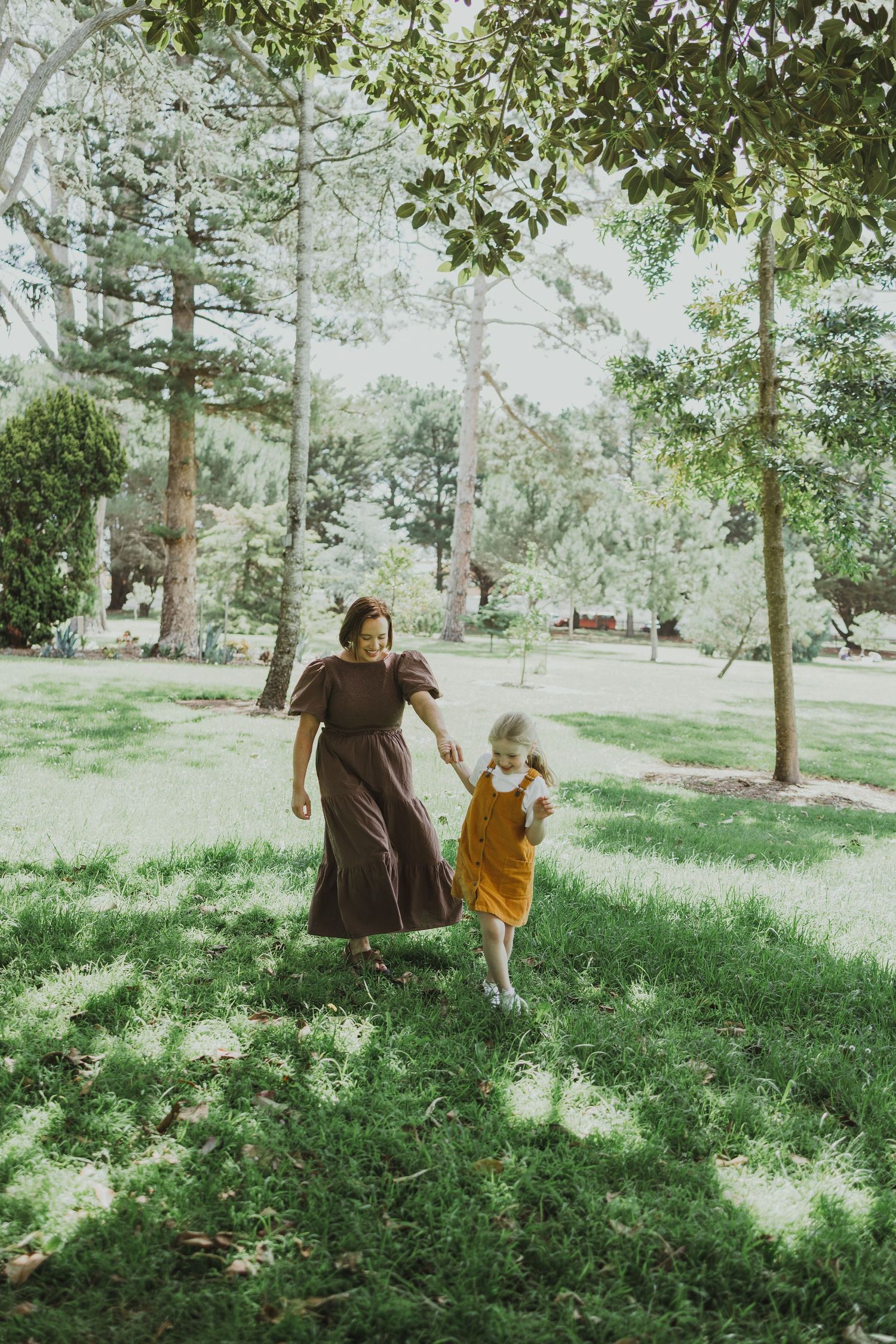 A mother in a brown dress holds her young daughter's hand while walking through a lush green park.