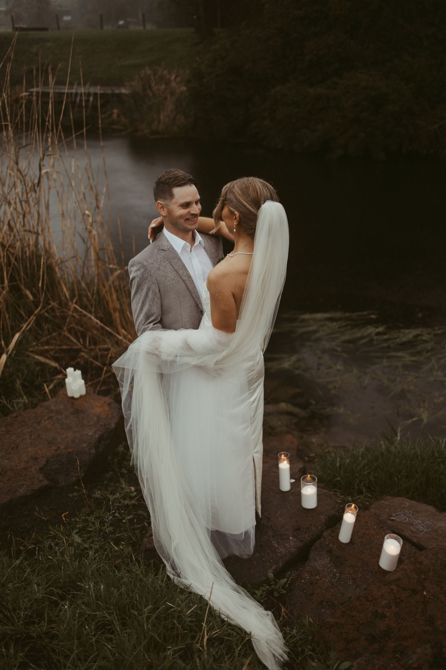 Romantic wedding couple embracing by candlelit stone steps near a misty pond, bride in white gown with veil.