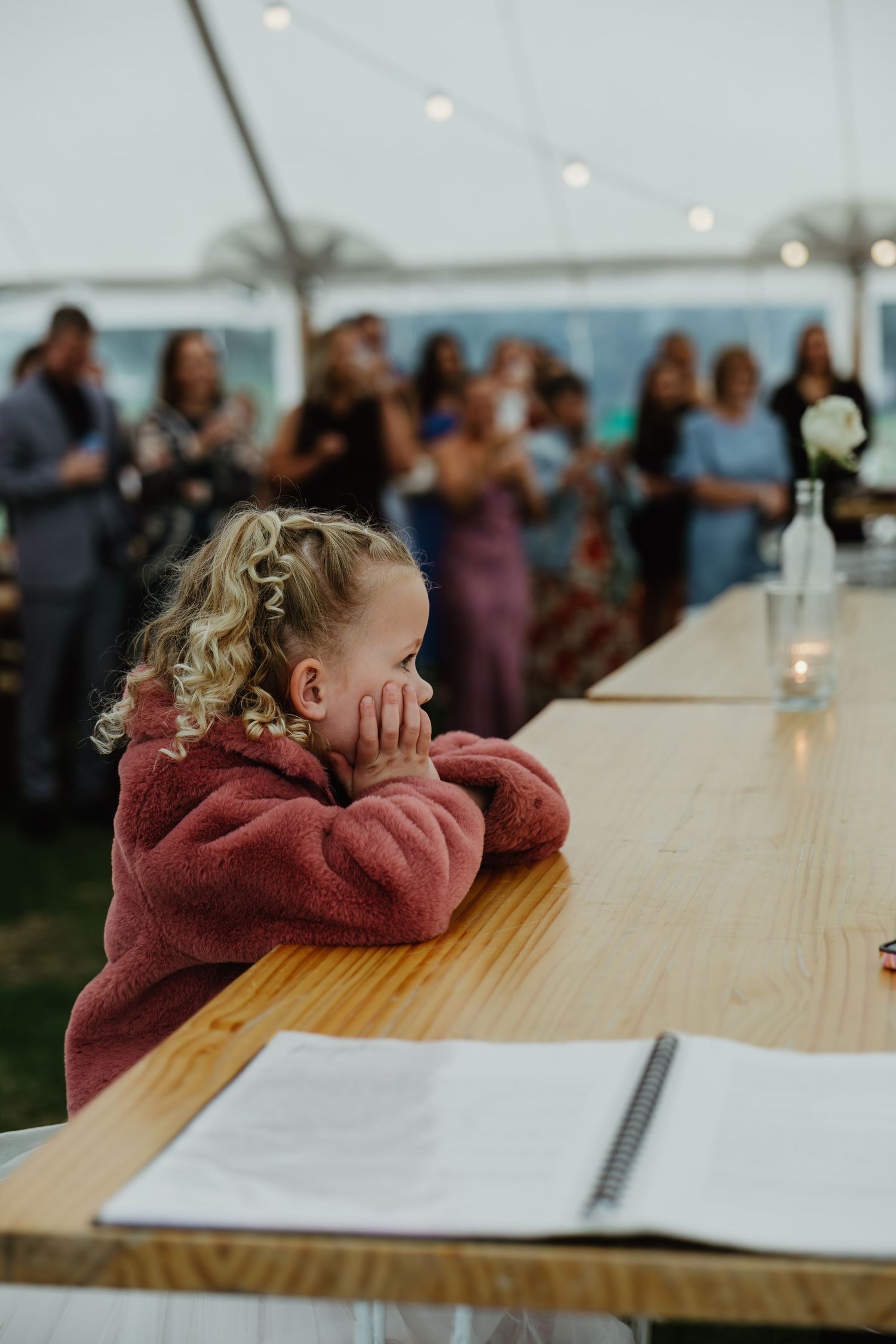Young curly-haired child in pink fleece resting at a wooden table inside a wedding marquee.