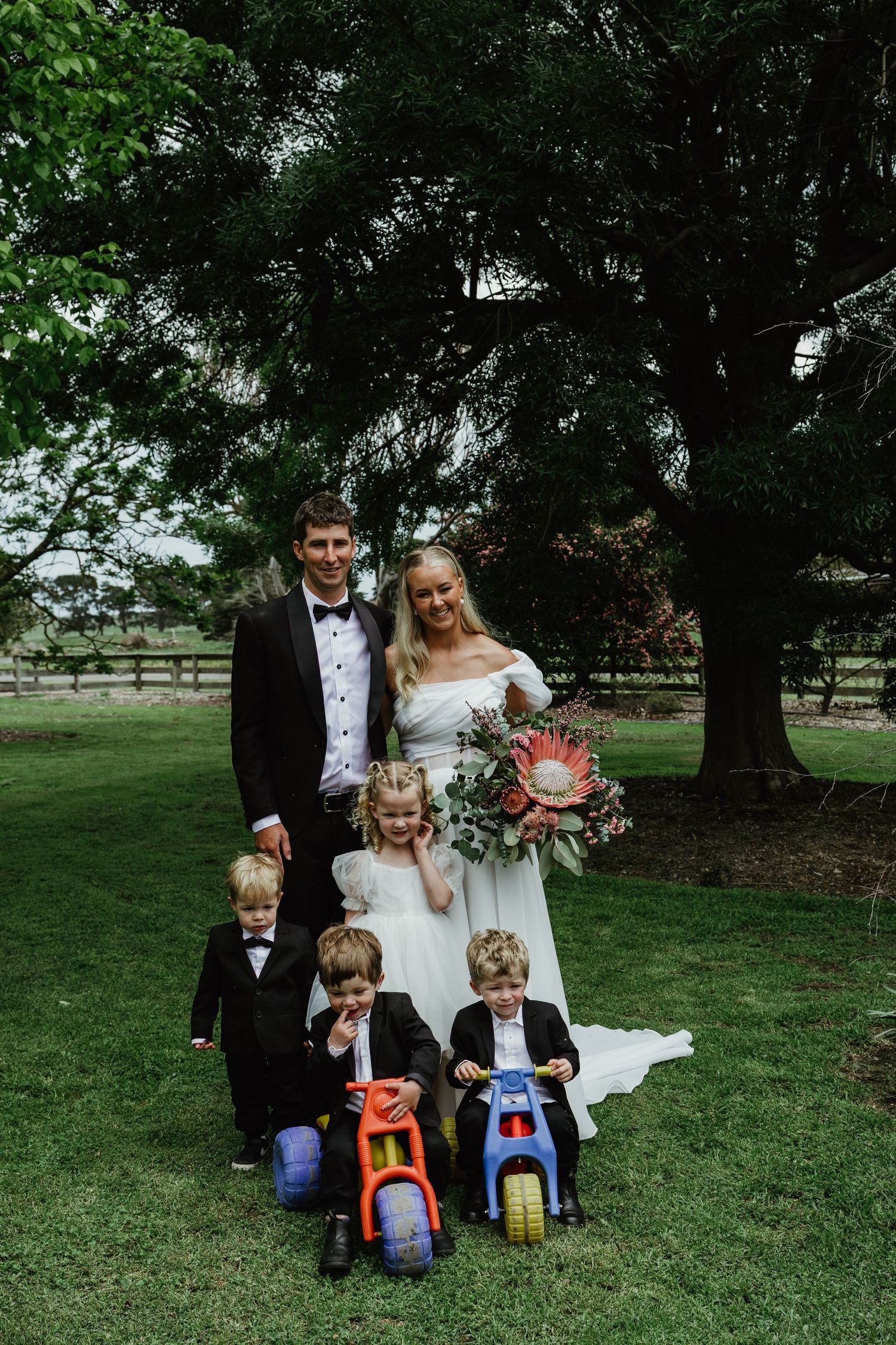 Wedding couple in formal attire posing with three children on toy bikes under large green trees.