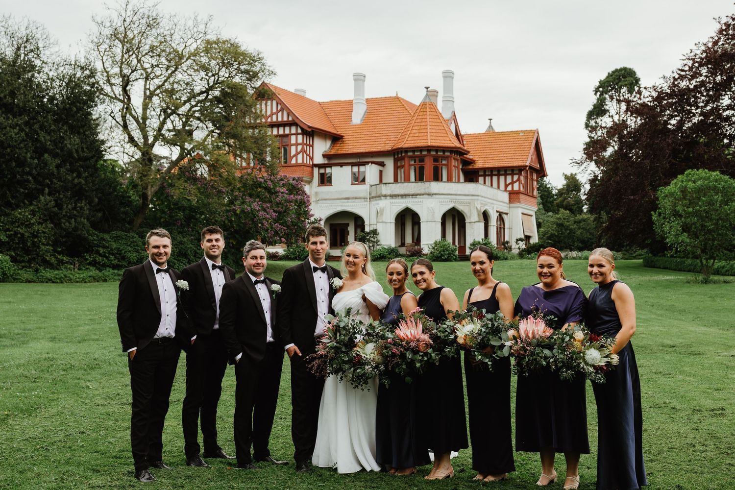 Wedding party poses before Tudor-style mansion with protea bouquets and navy dresses.