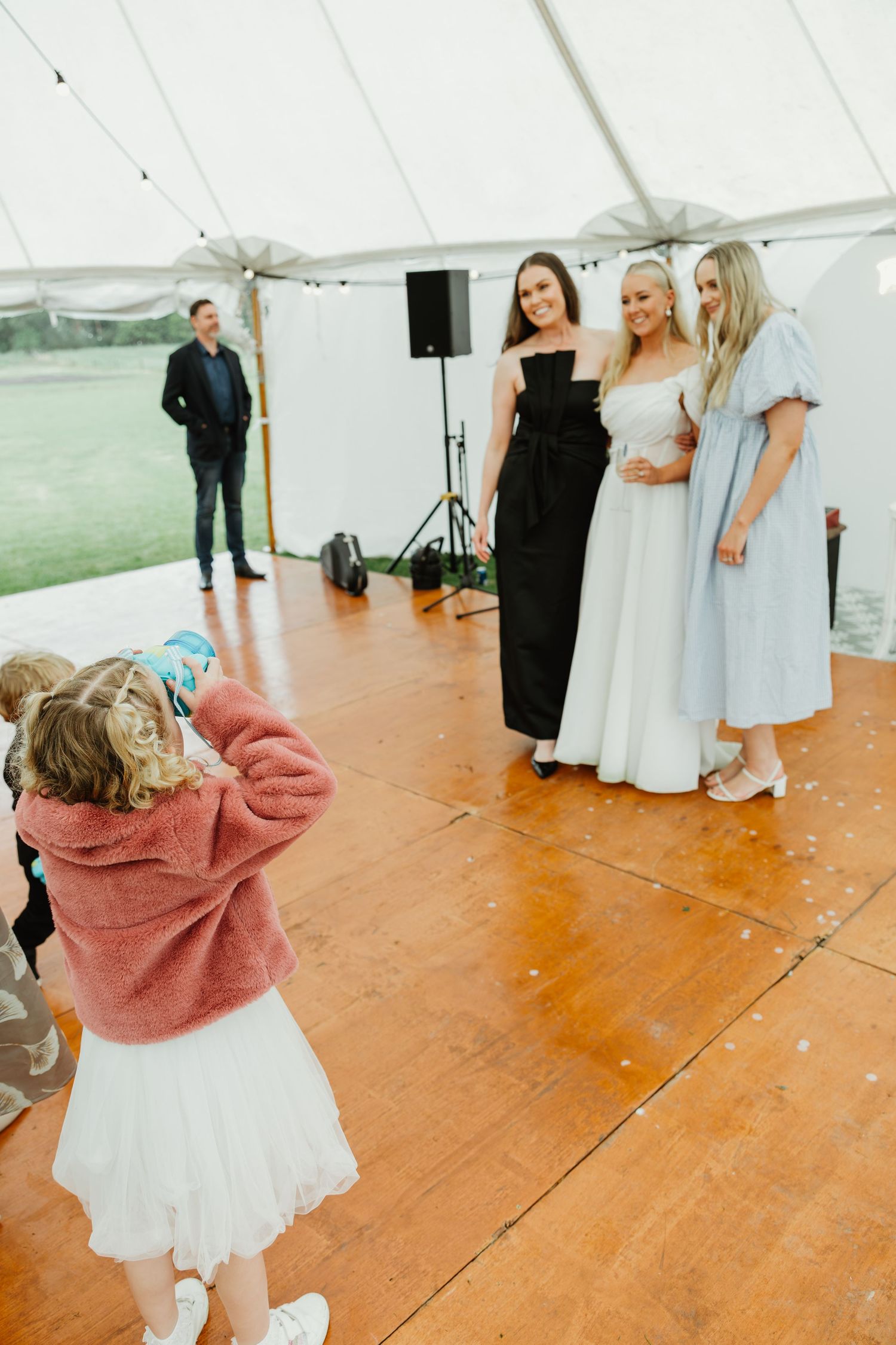 Young girl photographing bride and two friends posing together on dance floor inside wedding tent.