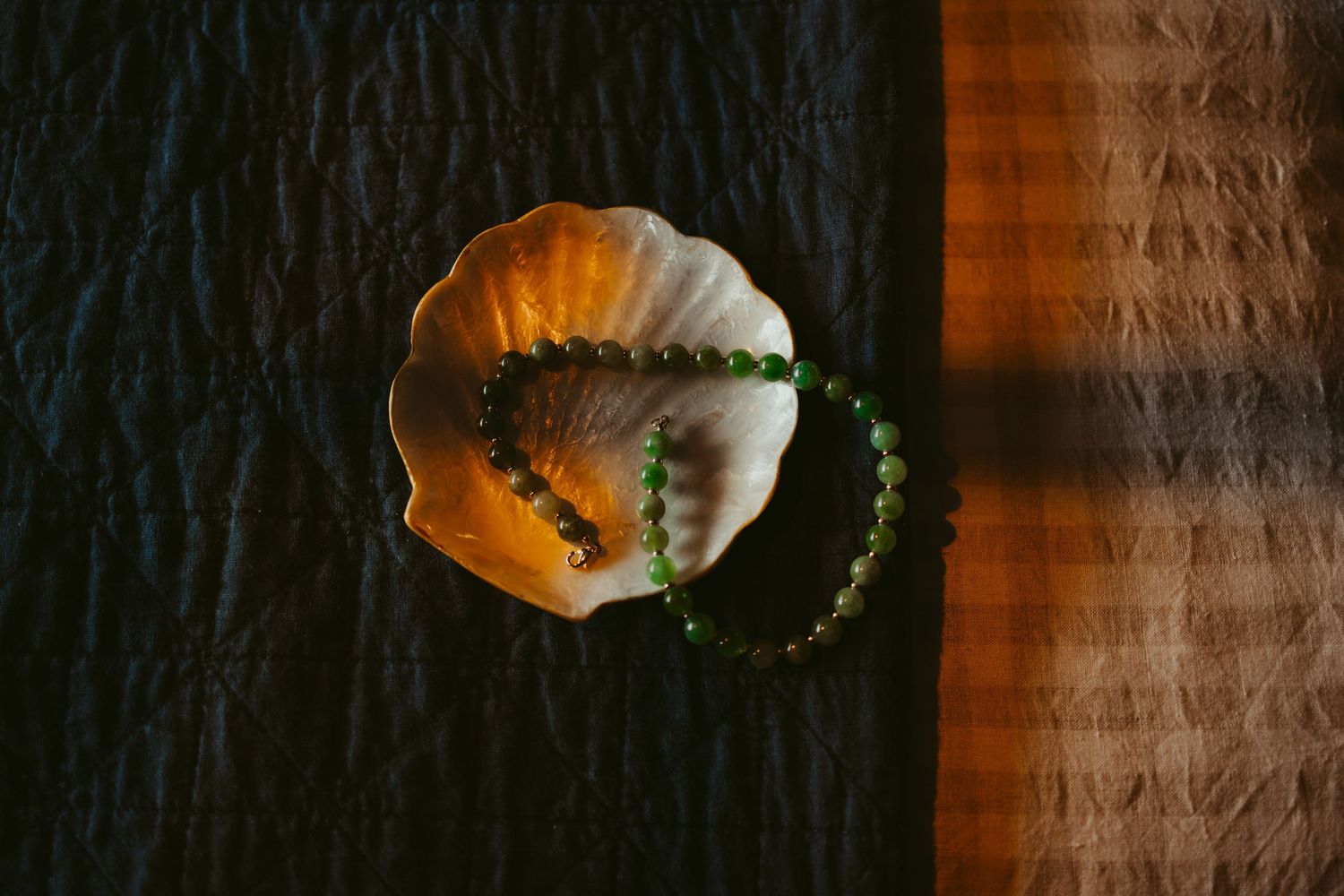 Green beaded bracelets displayed on a white shell dish, placed on a dark textured surface with warm light.