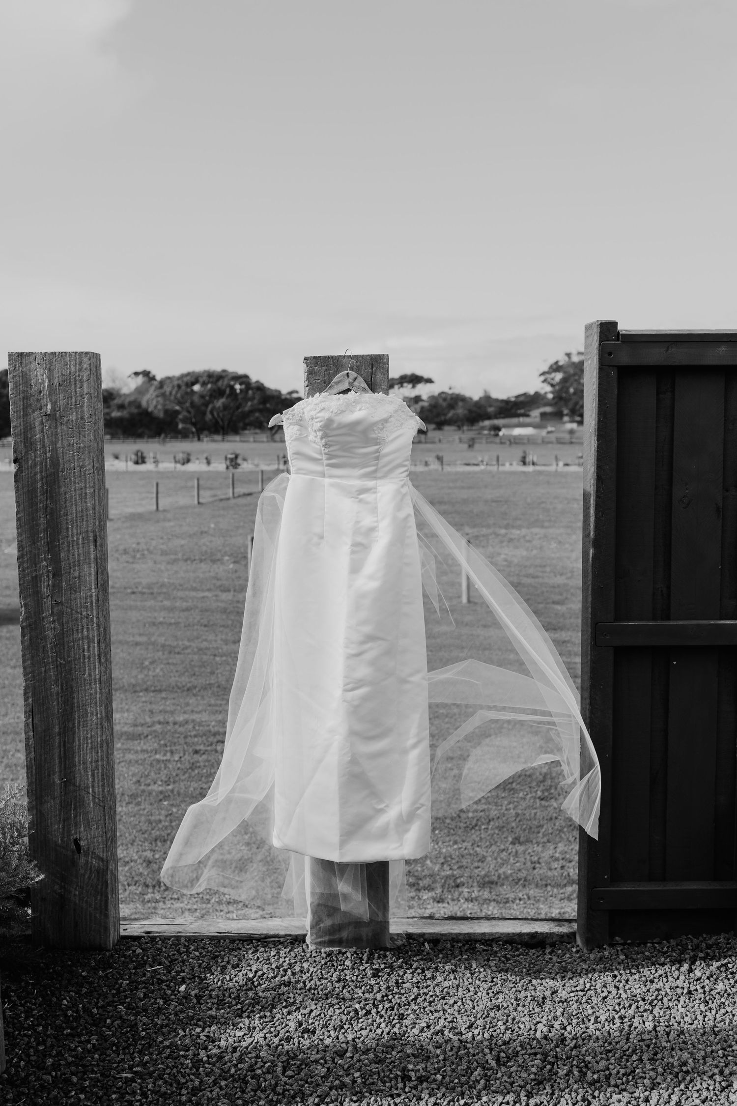 Elegant strapless wedding dress with flowing veil hanging outdoors between wooden posts on a farm property.