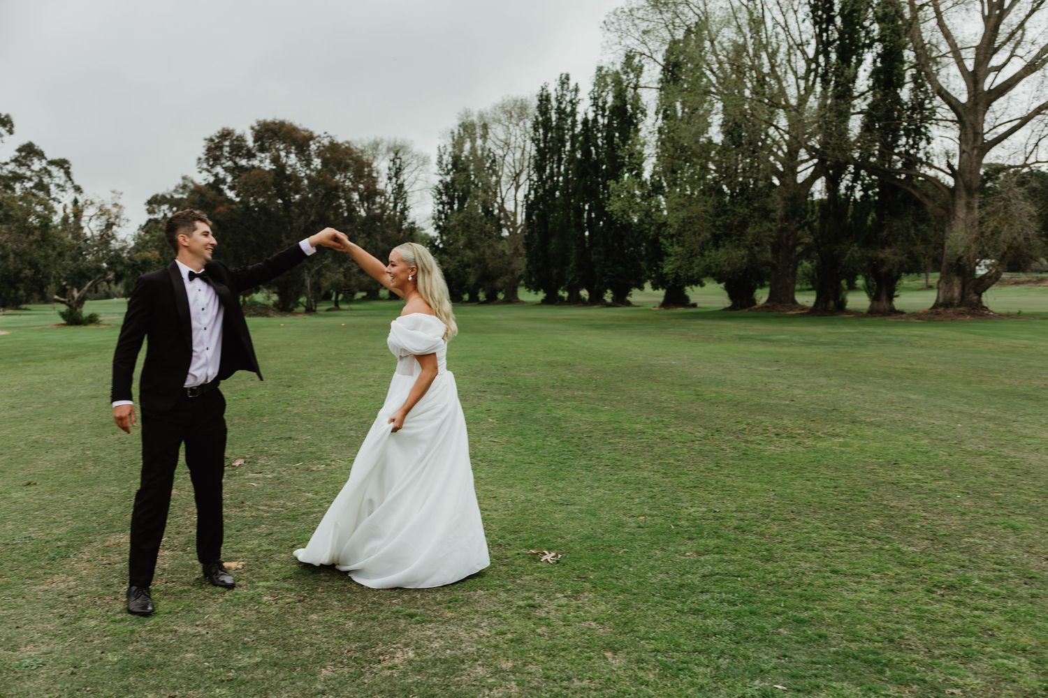 Bride and groom share joyful dance on lush green lawn with trees in background on wedding day.
