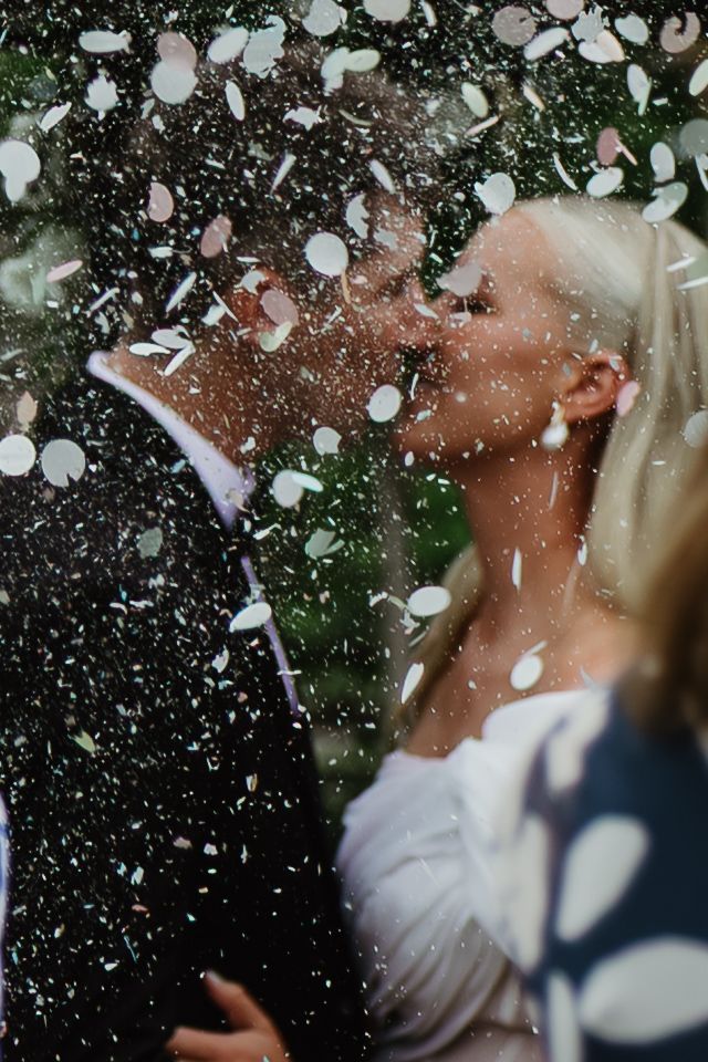 A bride and groom share a romantic kiss surrounded by falling white confetti petals at their wedding celebration.