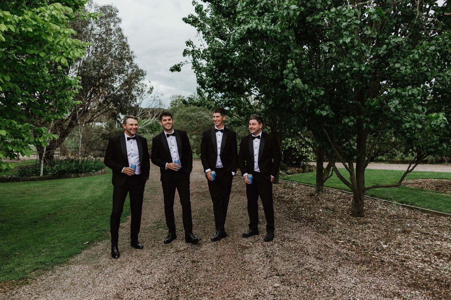 Four groomsmen in black tuxedos and bow ties pose outdoors in lush garden setting.