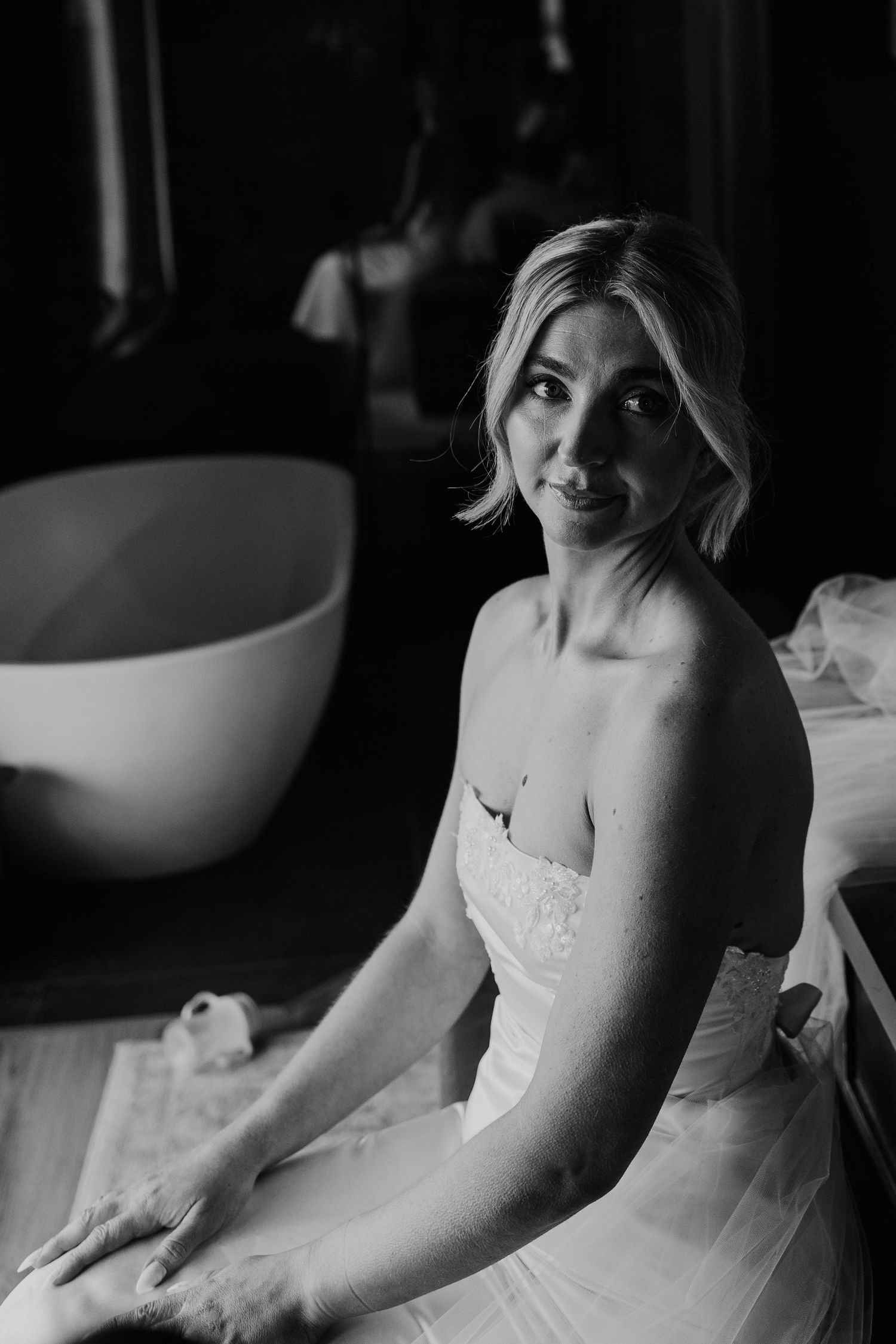 A bride in a strapless wedding gown sits pensively in a bathroom, captured in black and white.