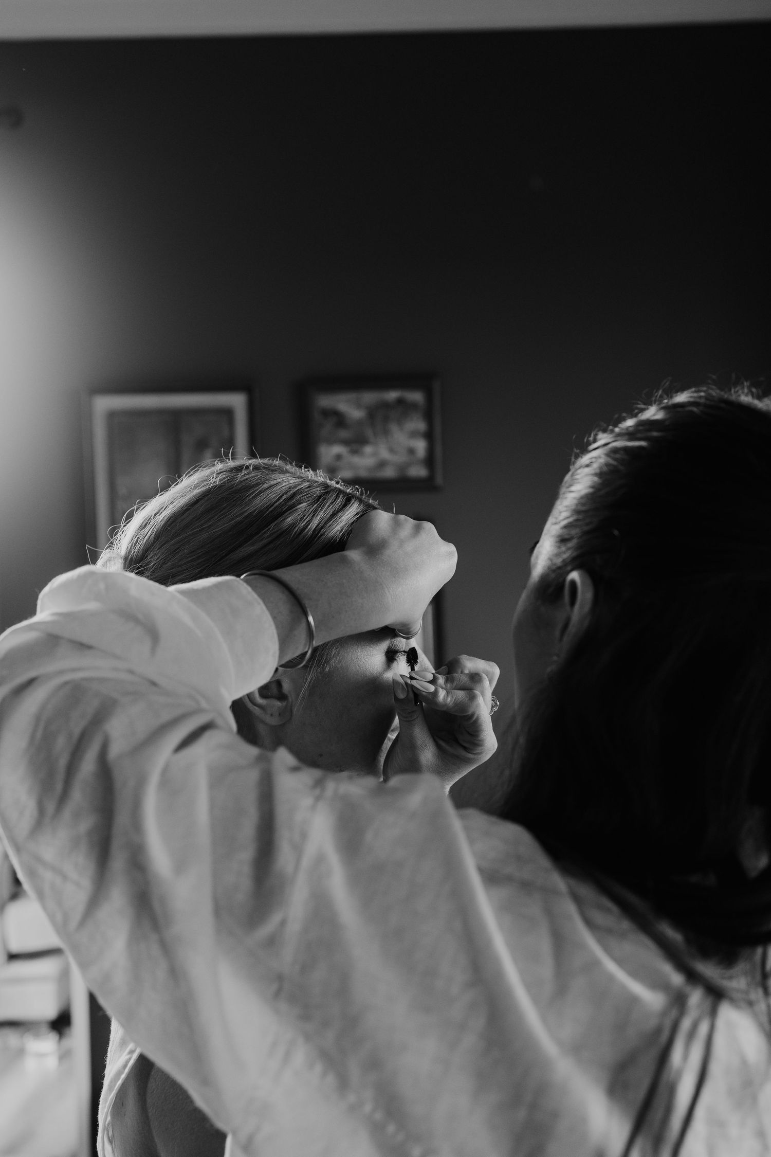 Black and white photo of makeup artist applying cosmetics to bride in white robe before wedding.