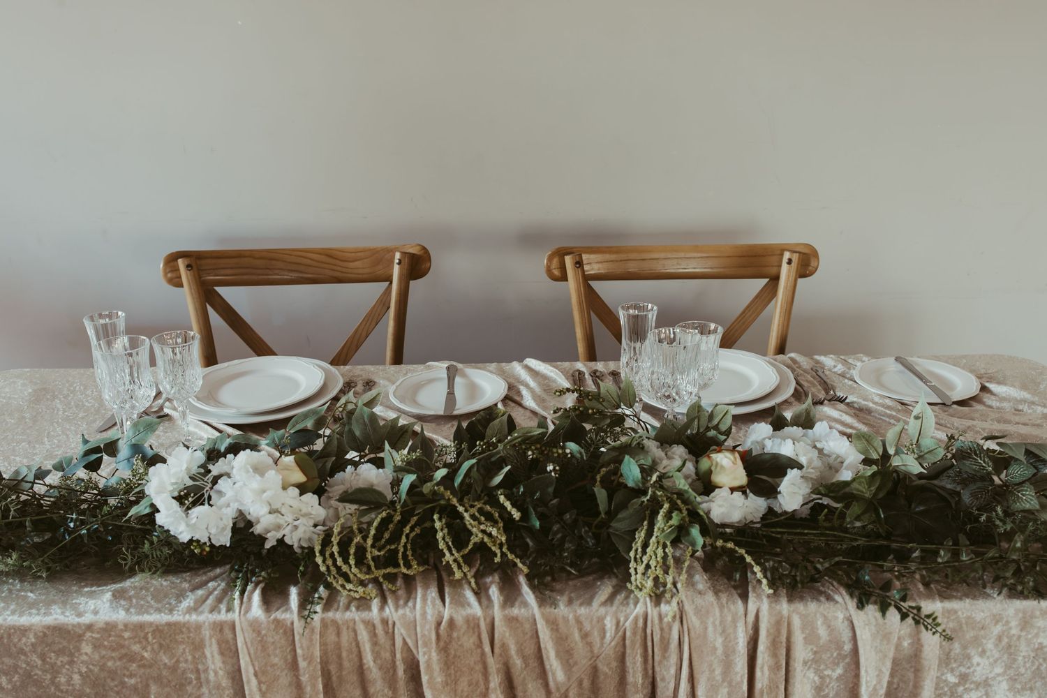 Elegant wedding sweetheart table with wooden cross-back chairs, white floral greenery runner, and neutral place settings.