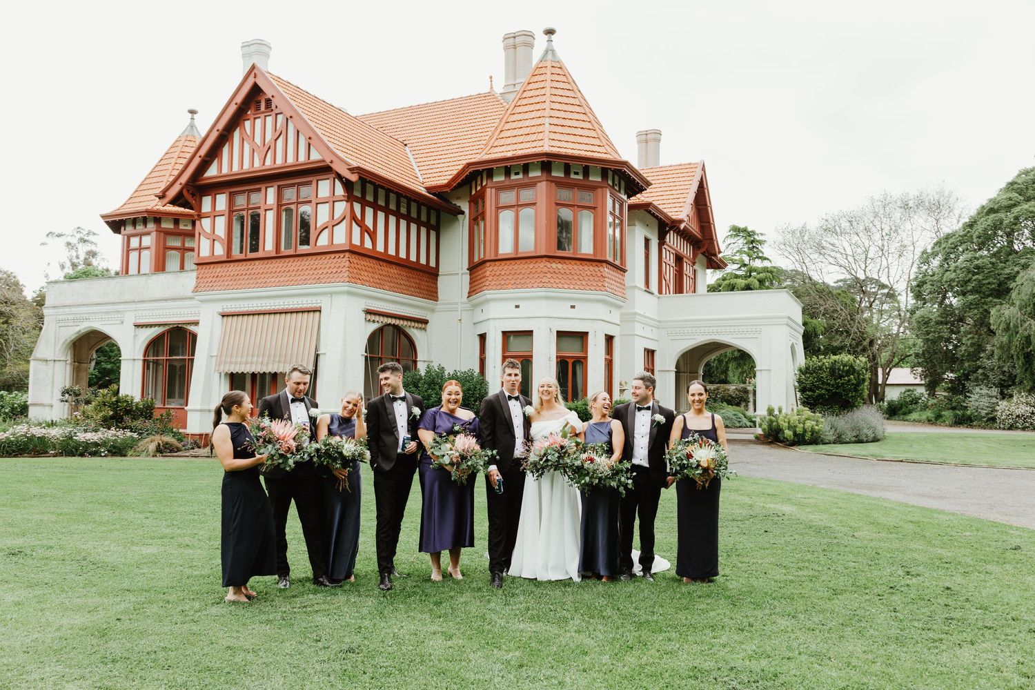 Wedding party in navy and black formal attire poses on lawn before elegant Tudor-style venue.