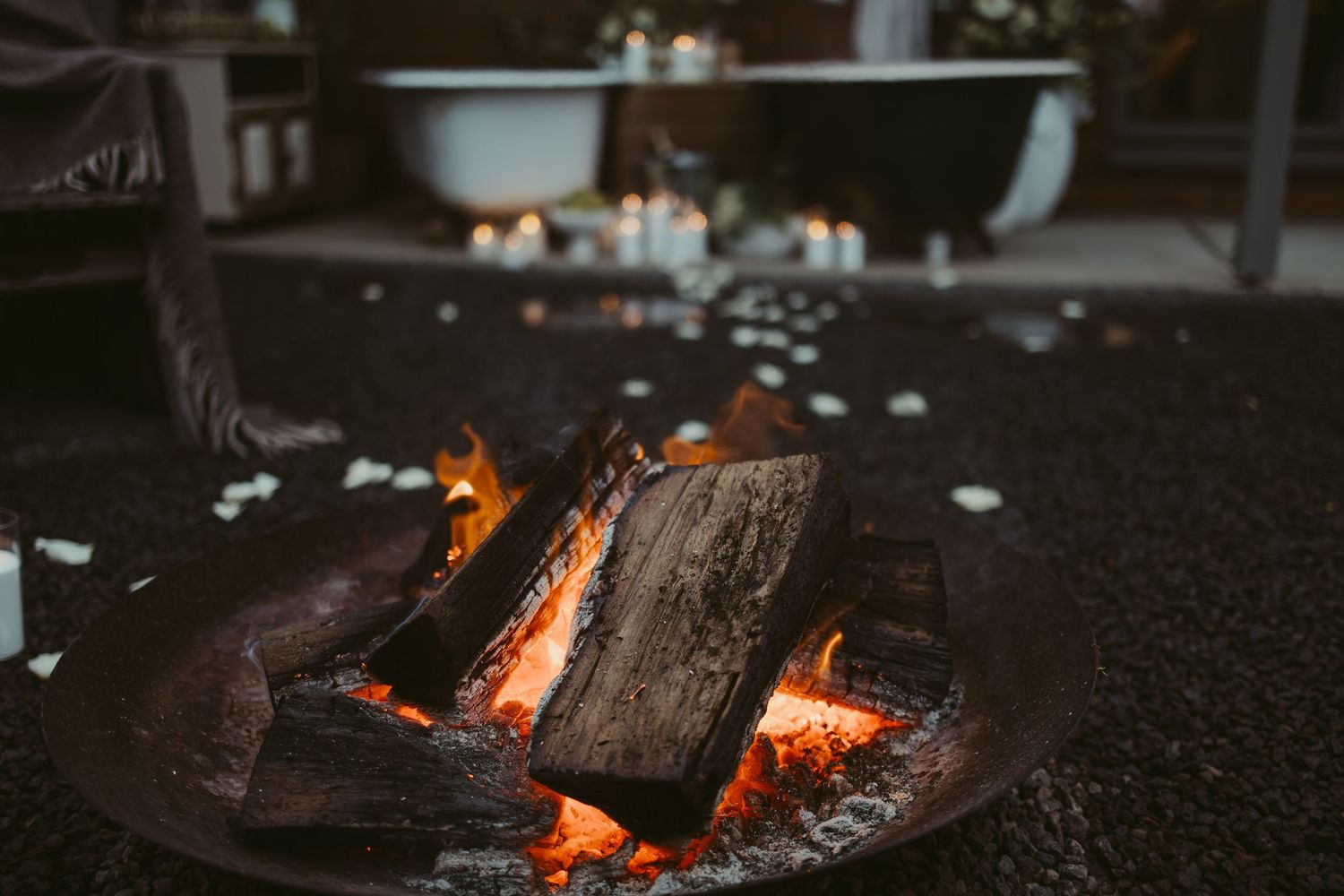 A rustic fire pit with glowing burning logs outdoors, with candles and a bathtub softly lit in the background.