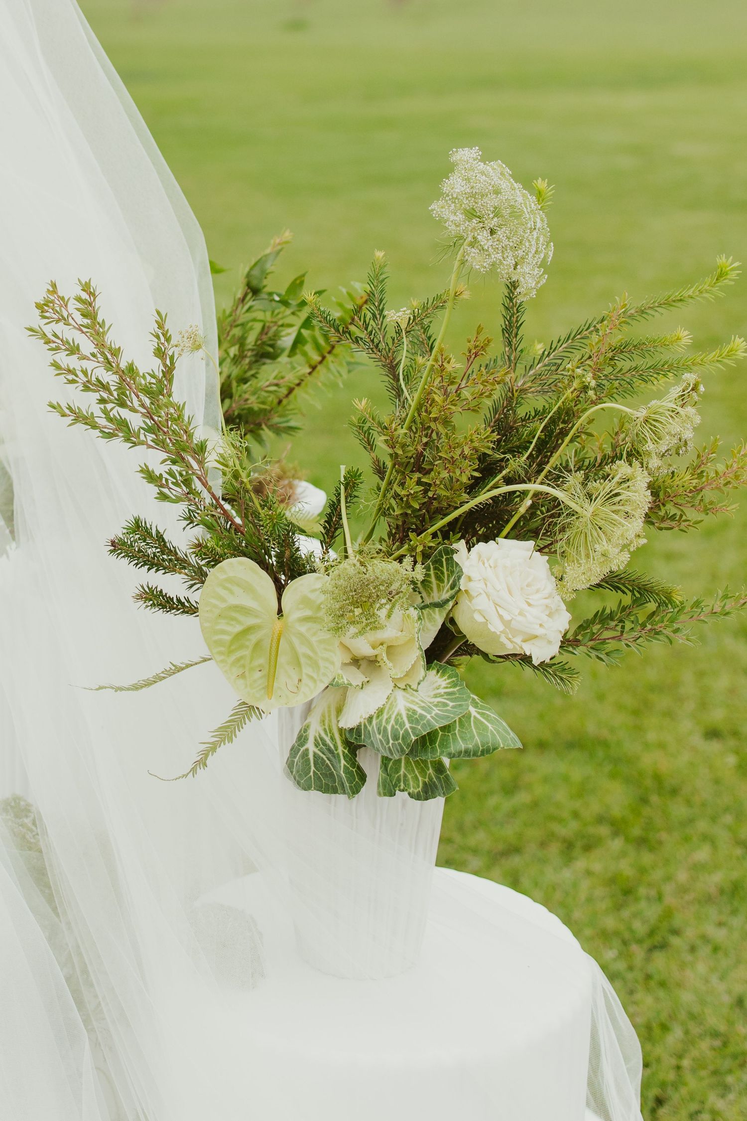 Lush white and green floral wedding arrangement in white vase on draped table against green grass background.