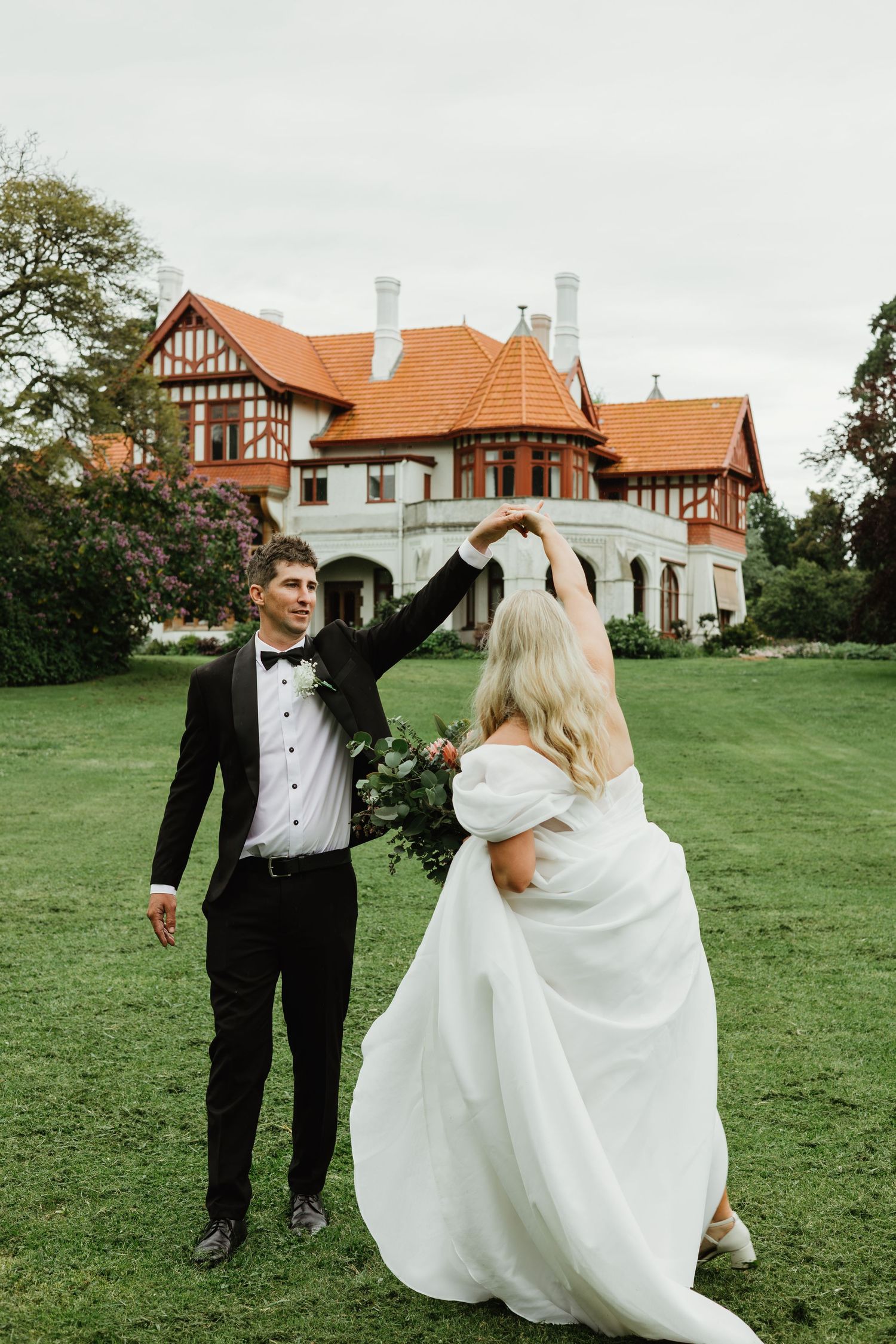 Groom spins bride in a white puff-sleeve gown on a green lawn outside a historic Tudor mansion.