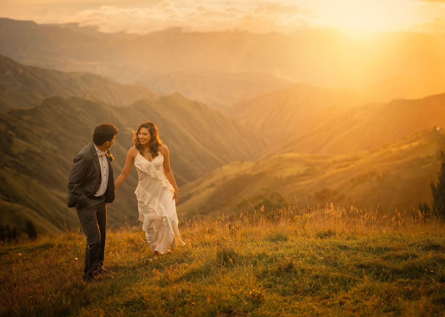 Couple in wedding attire stands on mountain ridge during golden sunset creating a romantic silhouette against the landscape.