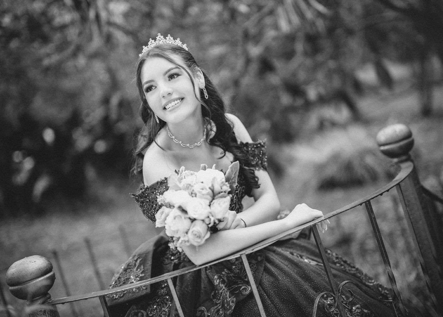 Black and white portrait photograph of a person sitting outdoors with flowers and a wooden bench in a garden setting.