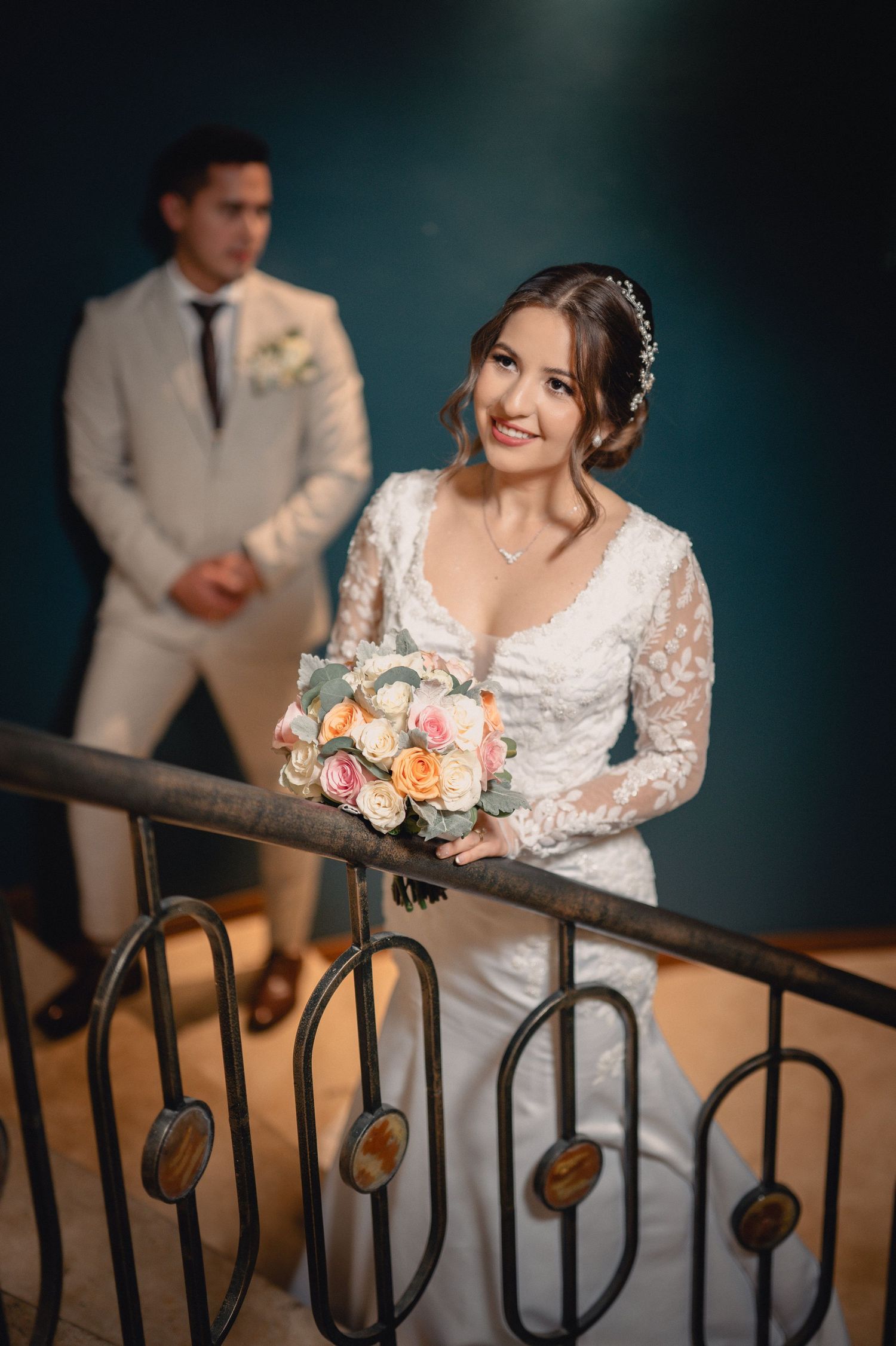 Wedding couple poses on a dark staircase with elegant white lace dress and peach floral bouquet in artistic sequence.