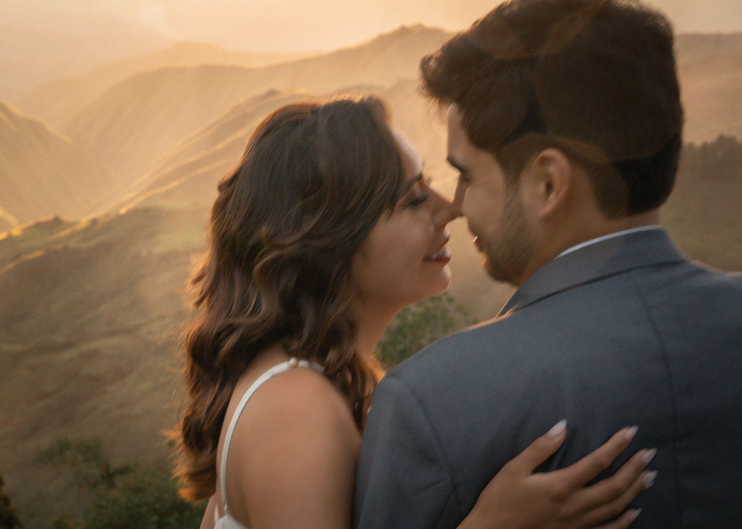 Romantic couple embraces on a mountaintop at sunset, silhouetted against a golden sky.