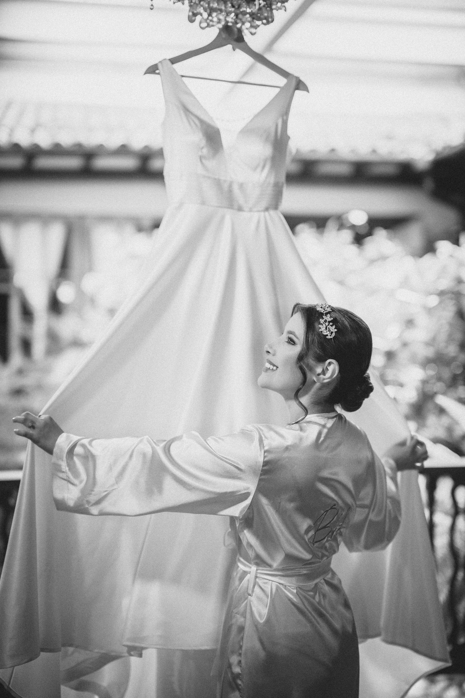 A classic white wedding dress hangs elegantly as someone in a silk robe reaches up to adjust it during preparation.