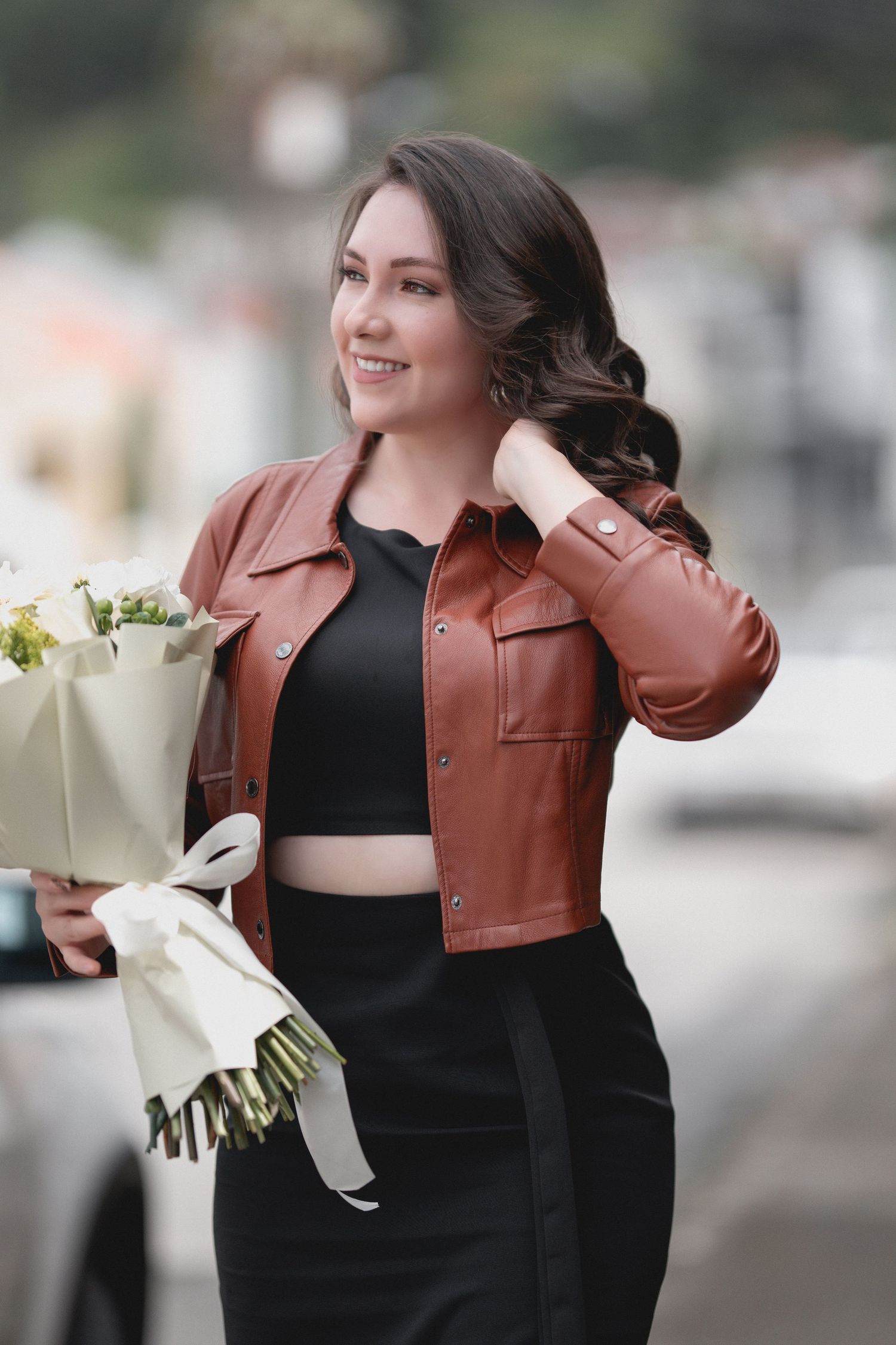 Fashion model wearing brown leather jacket and black dress poses with white flowers in urban setting.