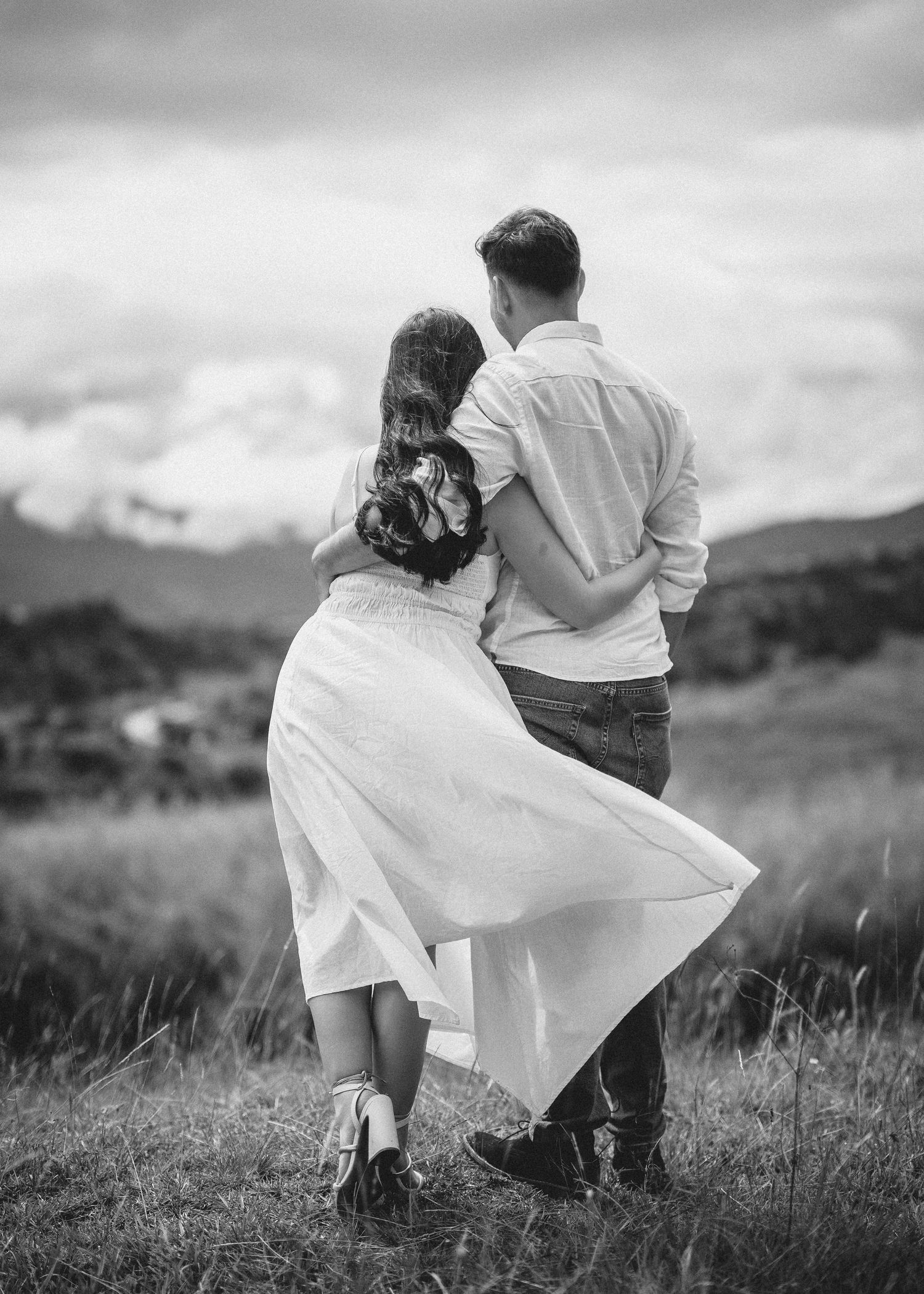 A romantic couple embraces in a windswept field with flowing white dress and dramatic black and white photography.