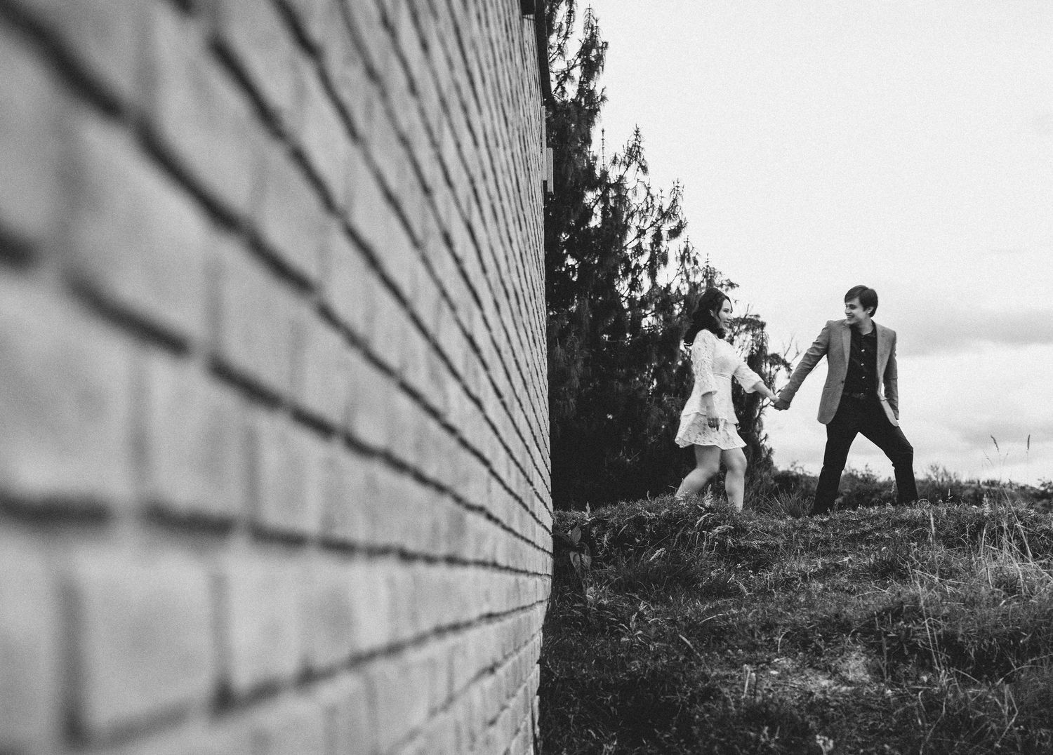 Black and white artistic photo showing two people running playfully along a brick wall in a natural outdoor setting.