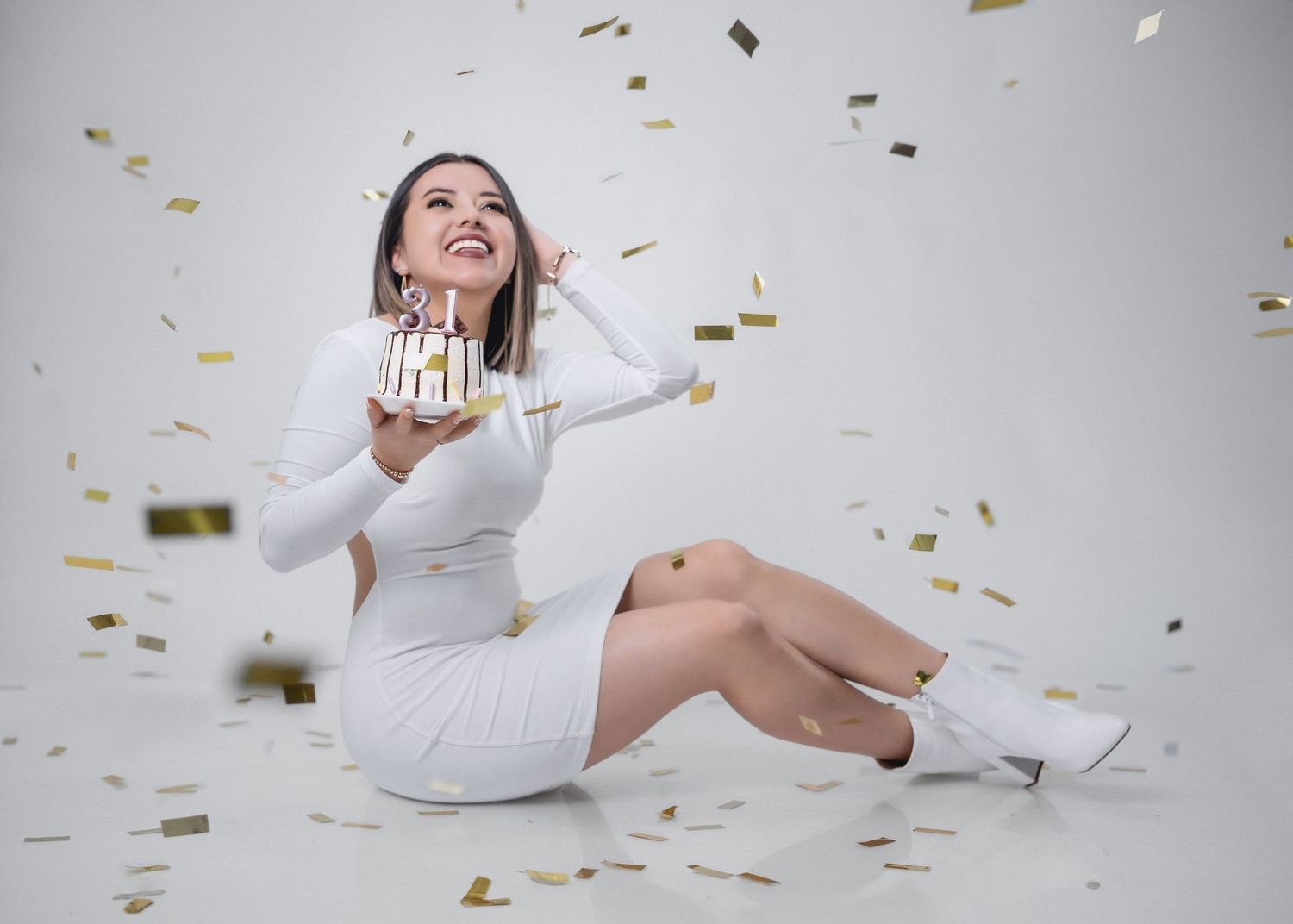 Woman in white dress lying on floor surrounded by falling gold confetti in celebratory photoshoot series.