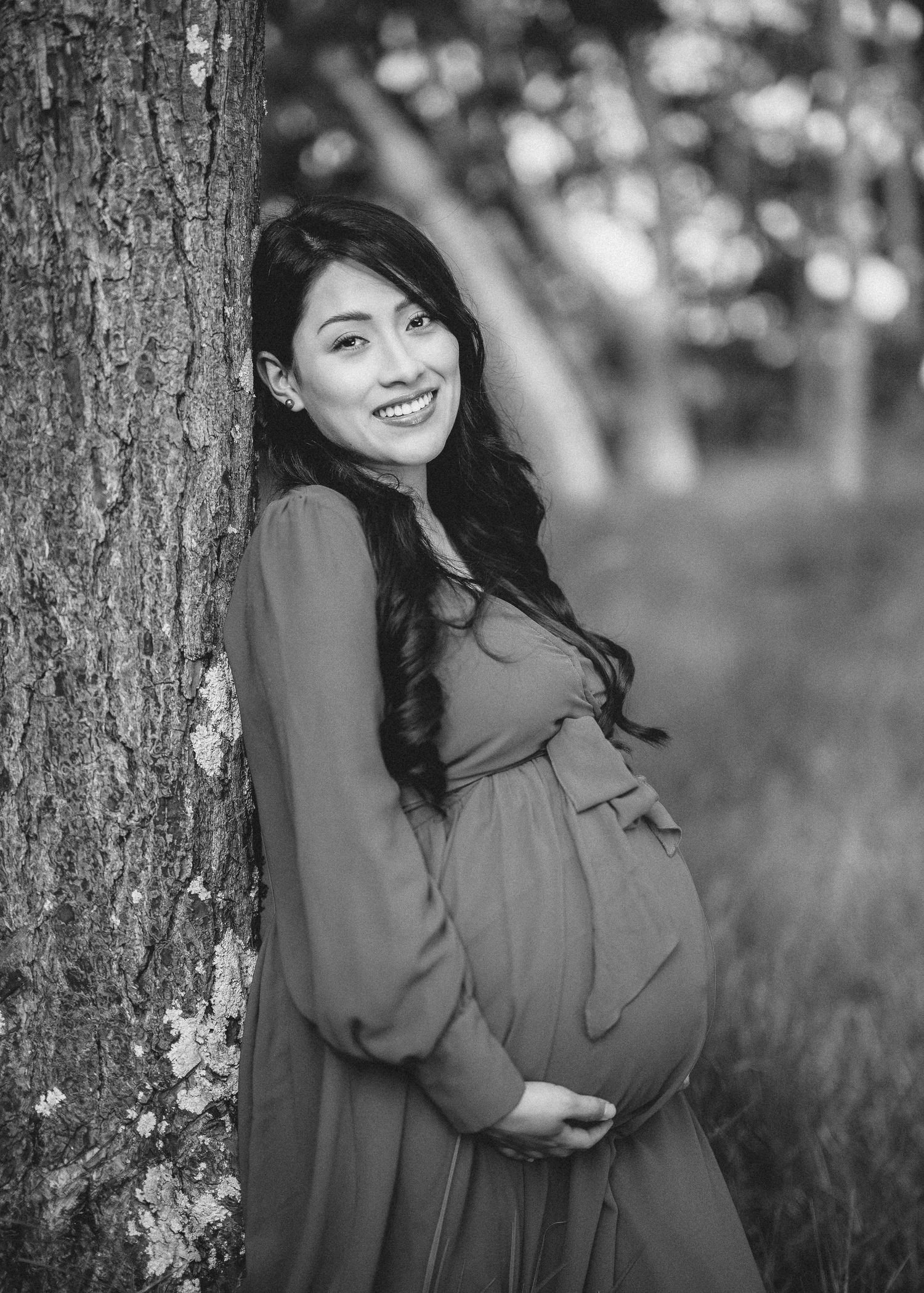 A person in a gray sweater poses against a tree trunk in a natural outdoor setting with bokeh background.