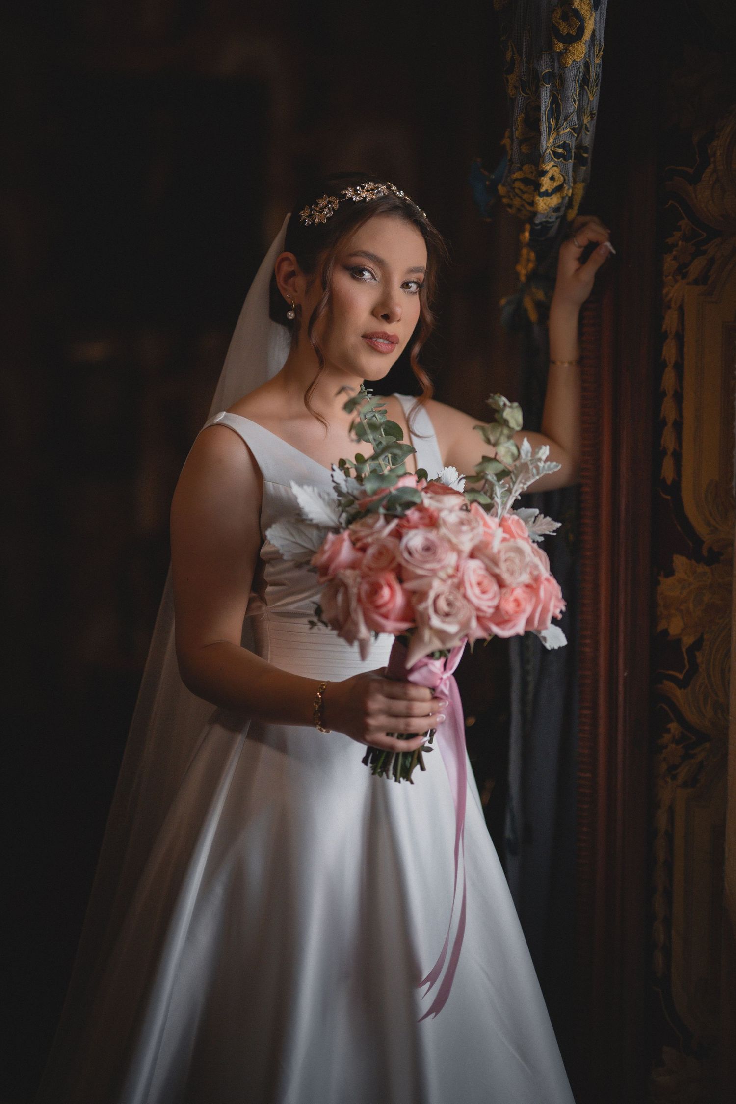 Classic bridal portrait featuring elegant white gown and pink rose bouquet against ornate dark background.