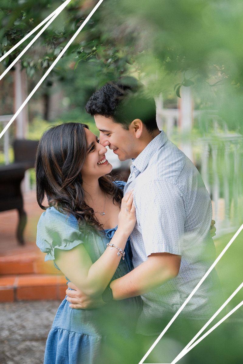 A romantic couple shares an intimate moment outdoors, embracing and smiling while framed by soft green foliage.