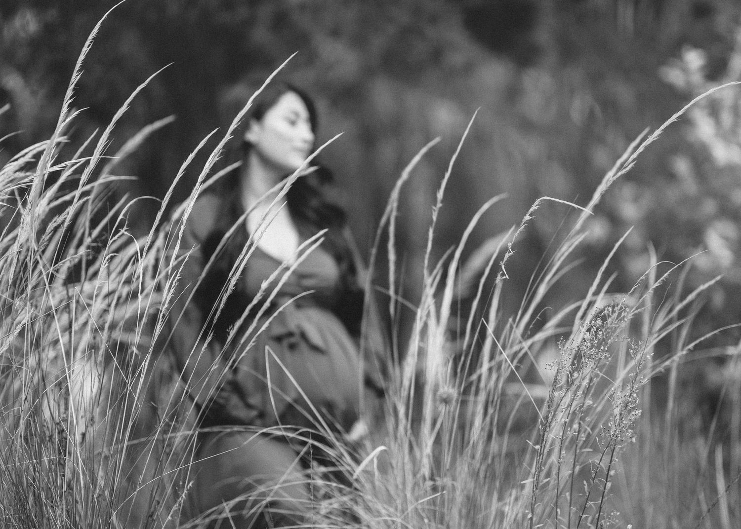 Black and white artistic portrait through tall grass showing figure in dark clothing against blurred natural background.