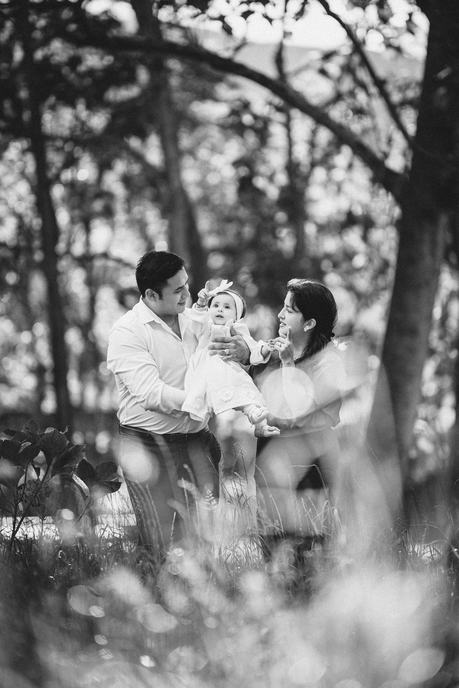 Black and white photo of a family playing together in a wooded outdoor setting with soft bokeh effects.