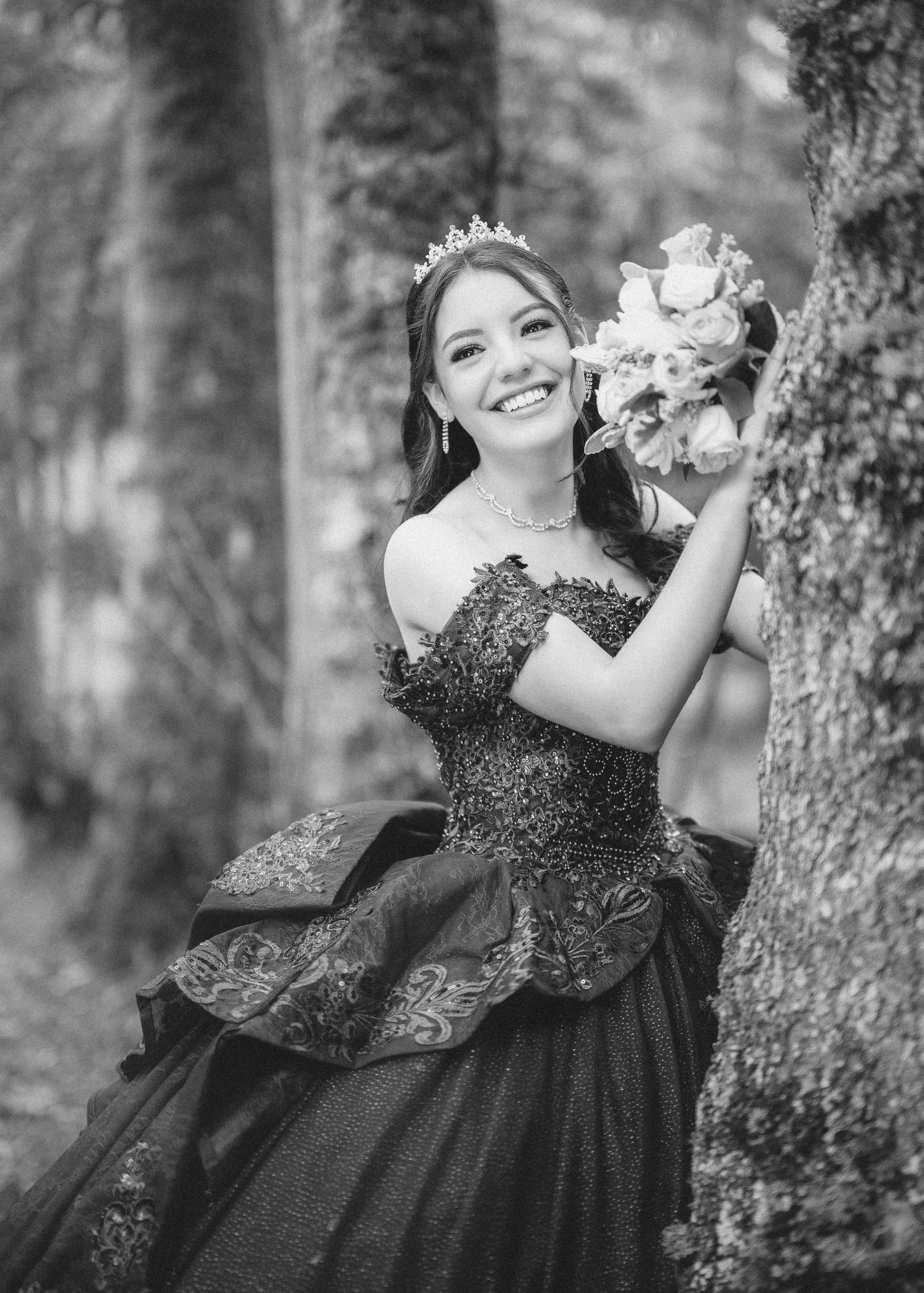 Person in a black formal dress poses gracefully against a tree trunk in a dreamy forest setting.